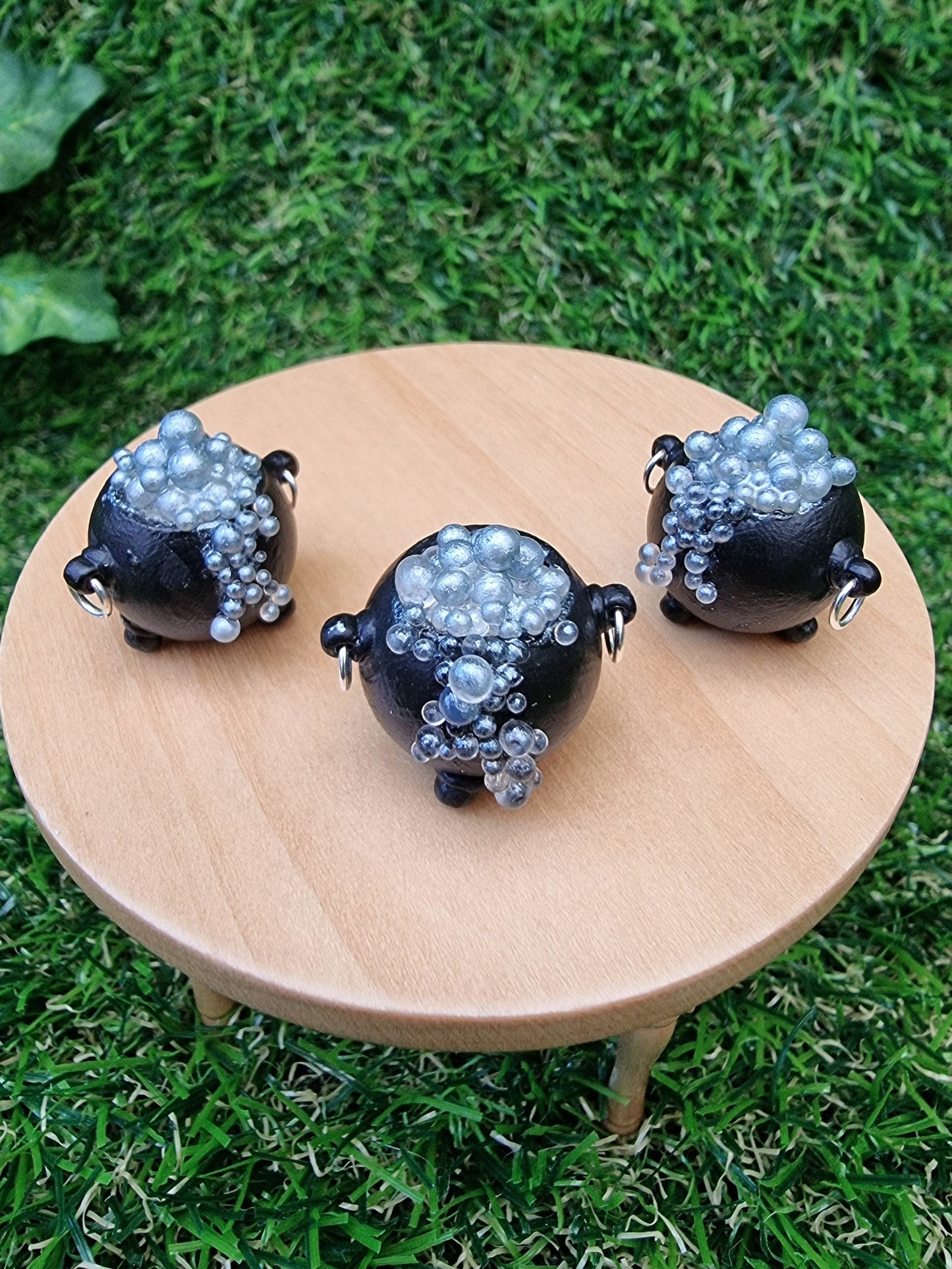 3 miniature black cauldrons overflowing with silver coloured bubbles are displayed on a miniature wooden table against a green grass background