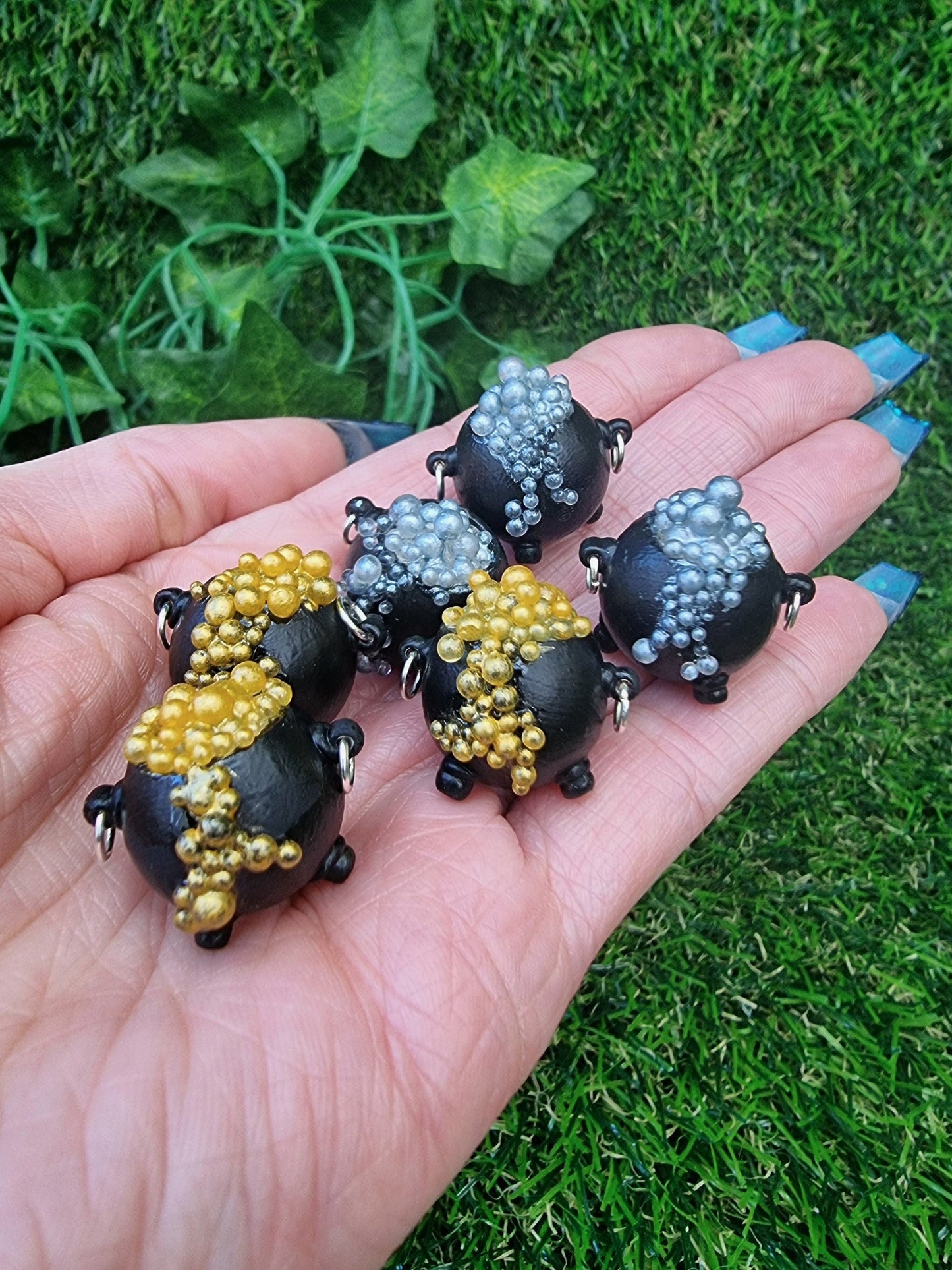 6 miniature black cauldrons overflowing with bubbles are displayed in an open palm. 3 of the cauldrons have bubbles in a gold colour. The other 3 have bubbles in silver colour
