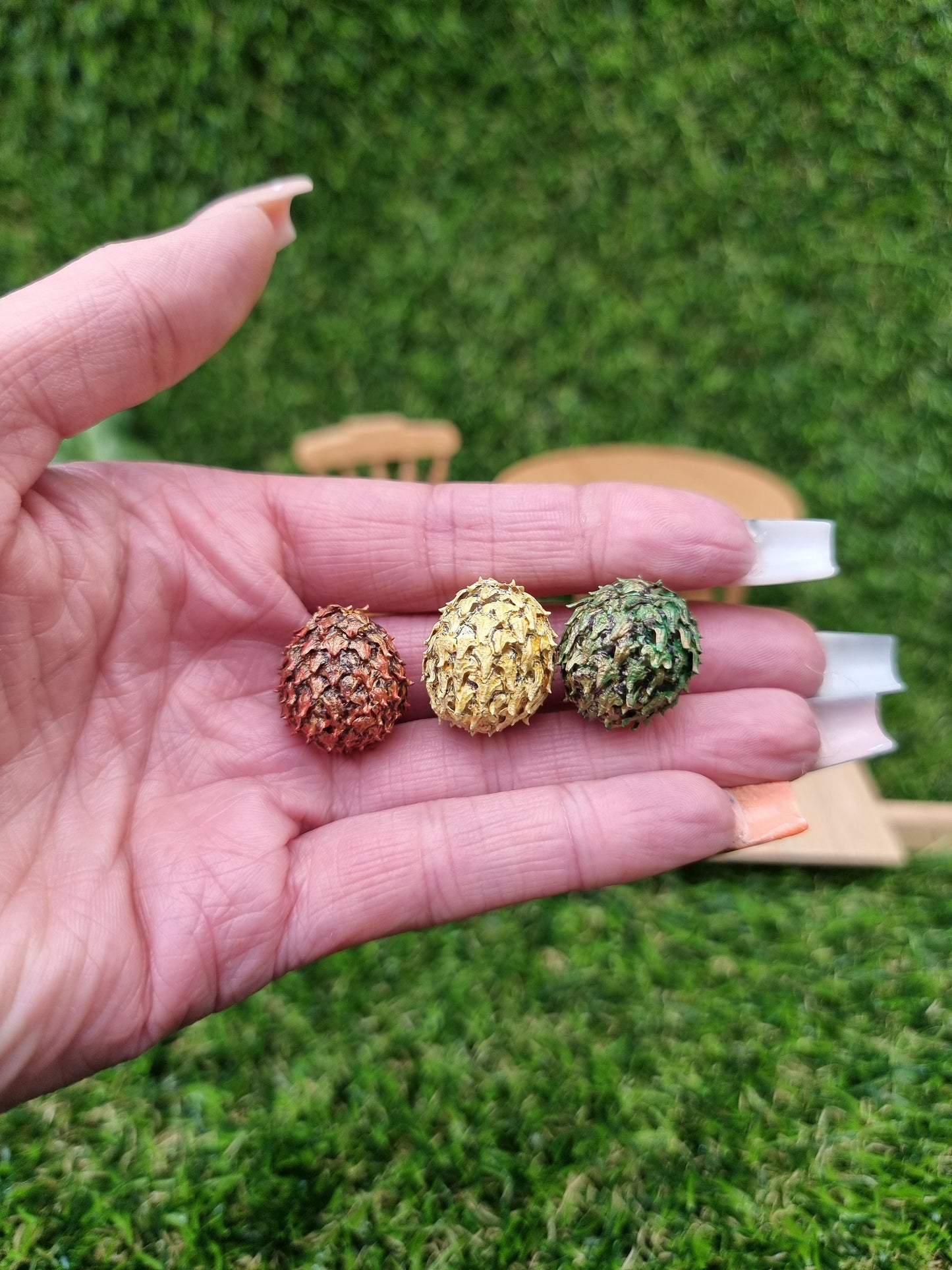 A hand holds 3 miniature scaled dragon eggs- one red, one green and one yellow, all with a gold sheen. Shown against a green background