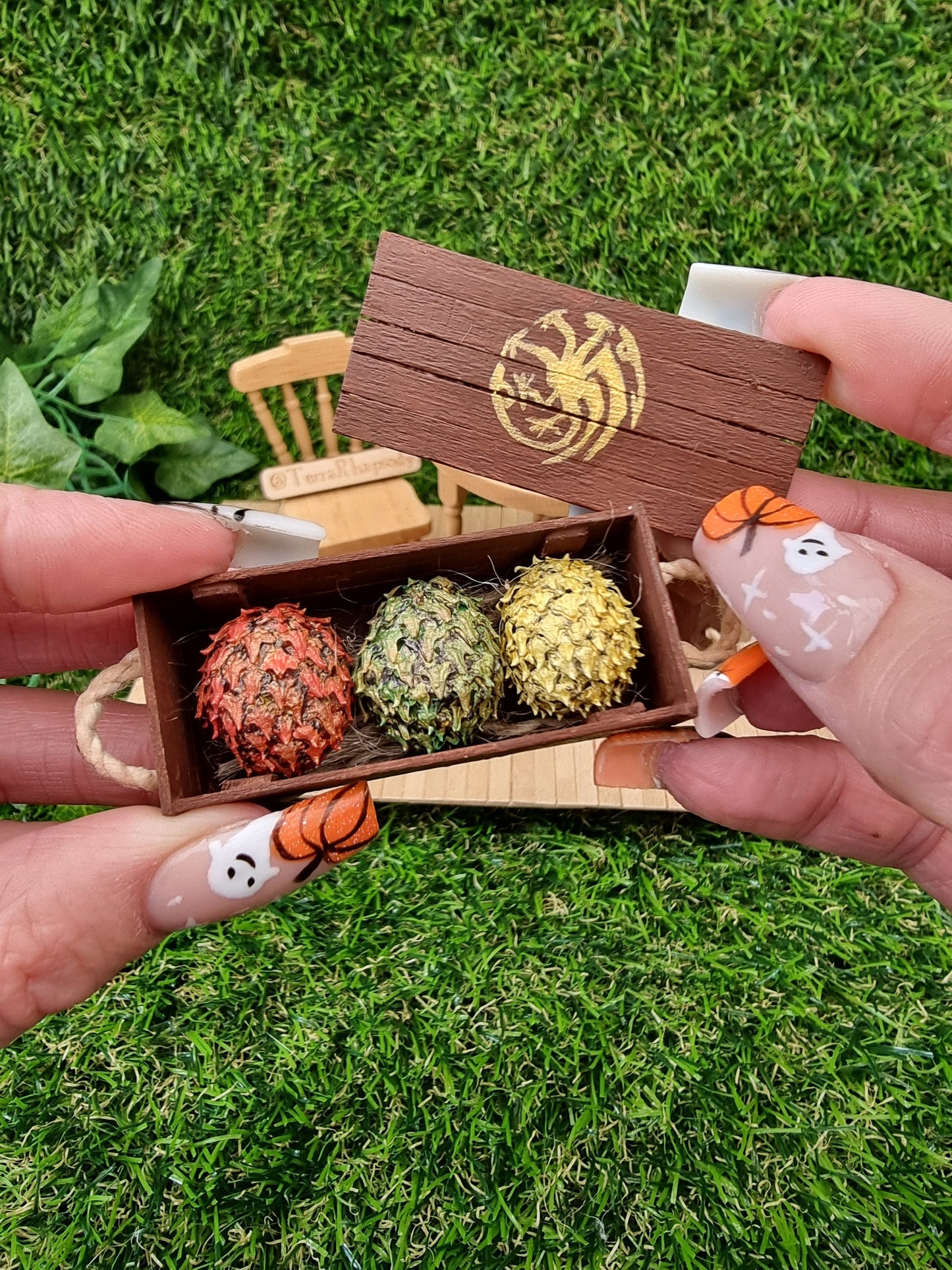 A hand holds a miniature brown crate and lid. The crate contains 3 scaled dragon eggs- one red, one green and one yellow, all with a gold sheen. The crate lid shows a gold Targaryen 3-headed dragon crest. The crate has rope handles oh the short edge