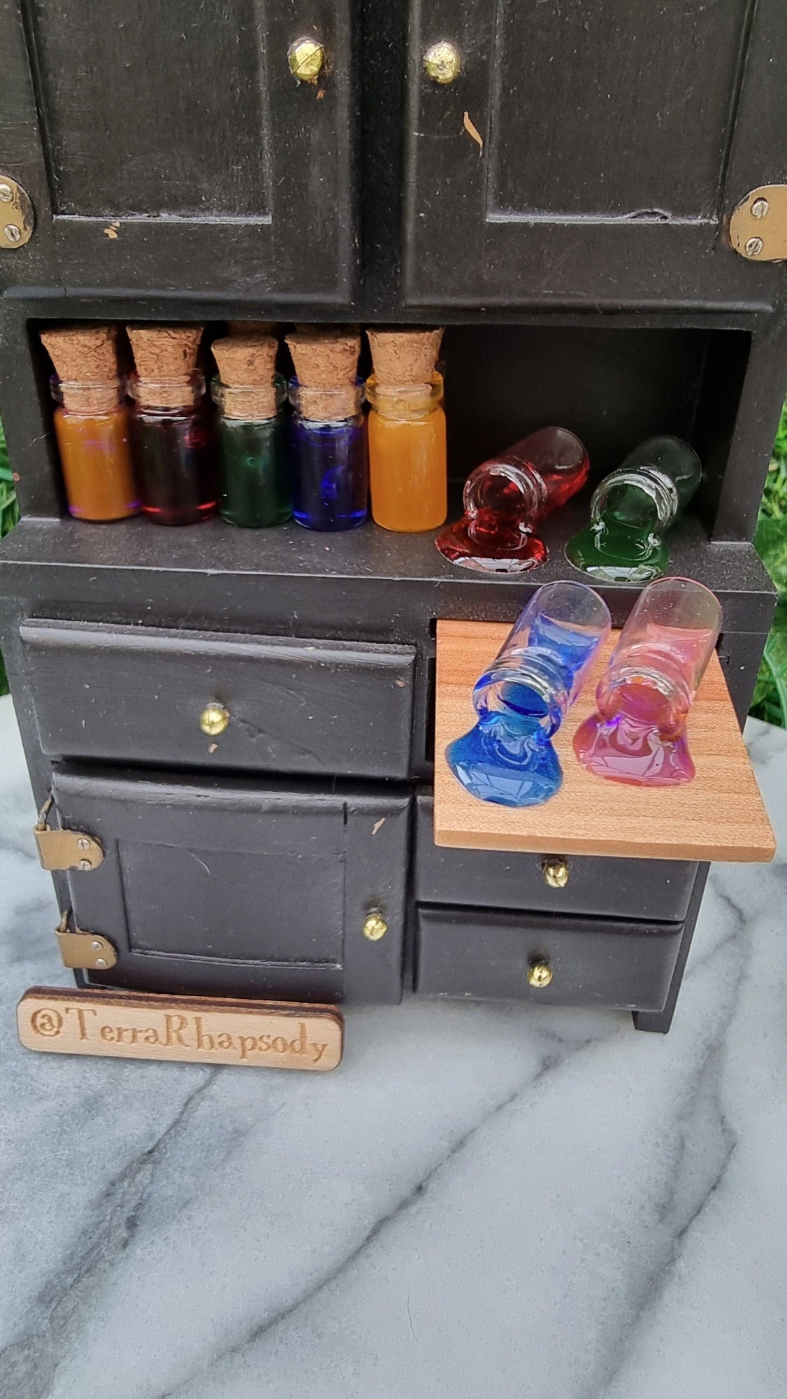 1:12 scale Miniature bottles with cork lids and different coloured resins are lined up on a miniature dresser. There are also some bottles on their side, appearing to have coloured potions pouring out of them