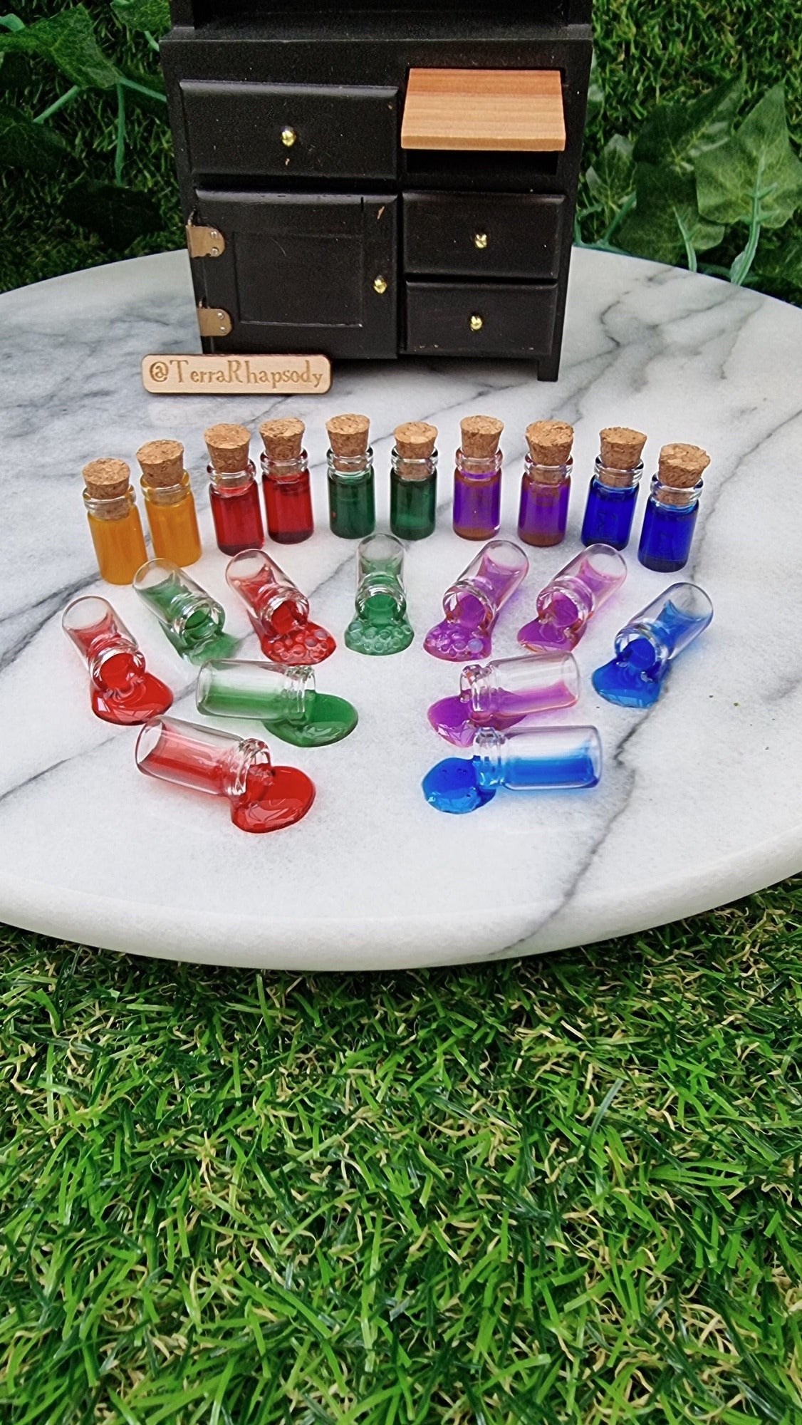 1:12 scale miniature potion bottles with cork lids and different coloured resins are lined up in front of a miniature dresser. In the foreground, a number of bottles are on their side appearing to have coloured with coloured potions pouring out of them. A mini black cabinet sits in the background and @TerraRhapsody is written on wood
