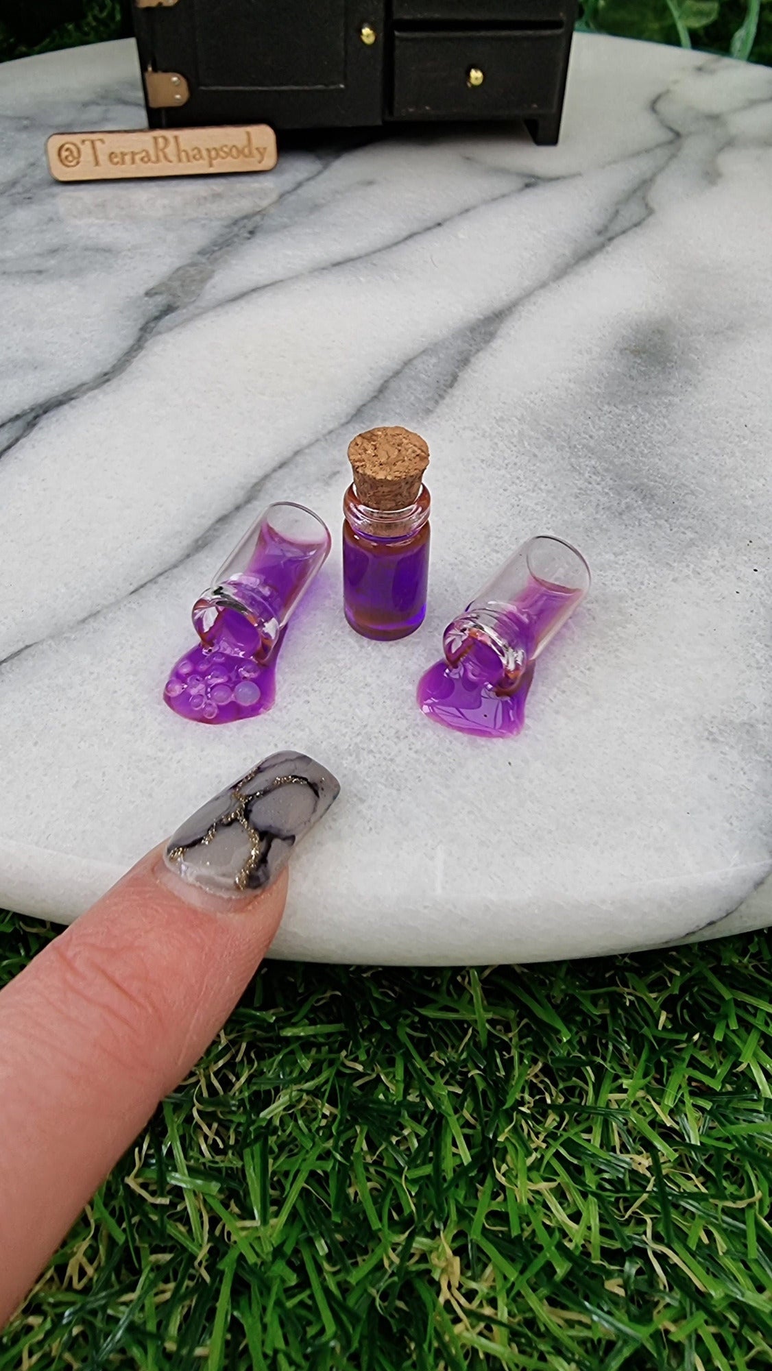 3 1:12 miniature bottles with purple potion are lined up in front of a miniature dresser. One bottle is filled and has a cork lid, 2 bottles are on their side appearing to have purple potion pouring out of them, 1 is bubbling. A finger points for scale