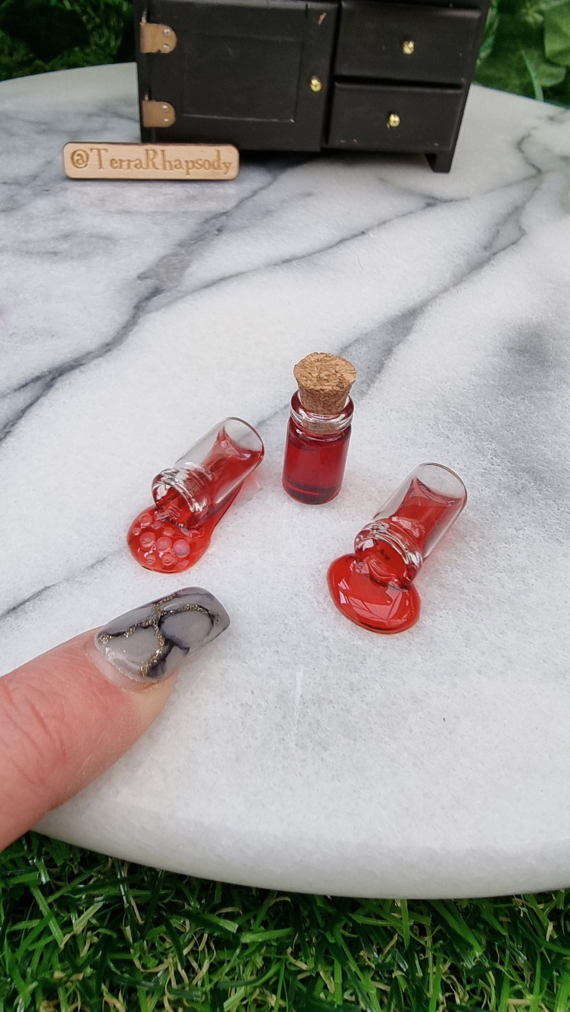 3 1:12 scale miniature bottles with red potion are lined up in front of a miniature dresser. One bottle is filled and has a cork lid, 2 bottles are on their side appearing to have red potion pouring out of them, 1 is bubbling. A finger points for scale