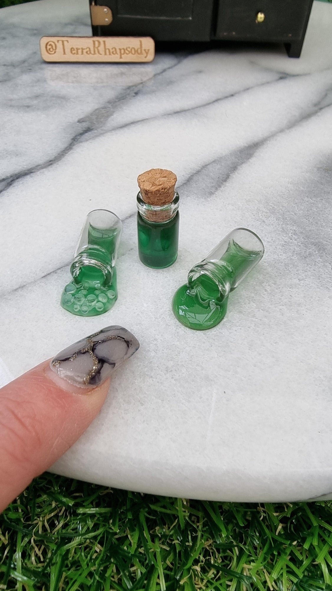 3 1:12 scale miniature bottles with green potion are lined up in front of a miniature dresser. One bottle is filled and has a cork lid, 2 bottles are on their side appearing to have green potion pouring out of them, 1 is bubbling. A finger points for scale