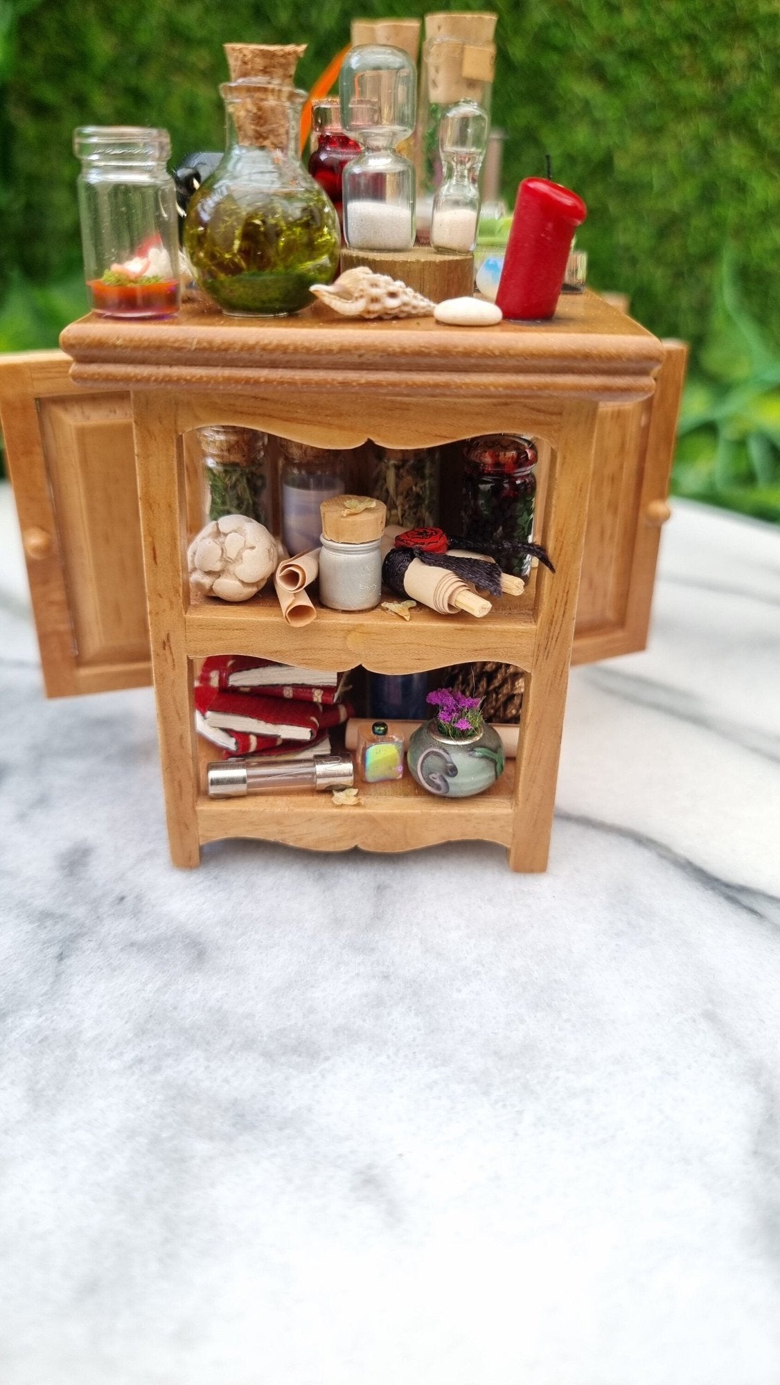 Side view of a 1:12 scale minature wooden sideboard showing open shelves at the end. The shelves contain parchment scroll, vase, glass tube, books curios and bottles. The top has a shell, candle, sandtimers, green potion and glass jar