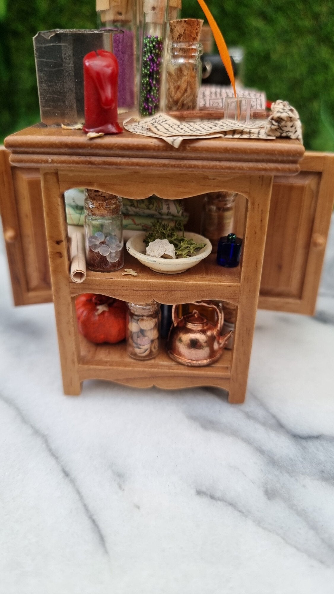 Side view of a 1:12 scale minature wooden sideboard showing open shelves at the end. The shelves contain map rolls, copper kettle, pumpkin, dish and bottles. The top has parchments, candle, filled bottles, inkwell and quill