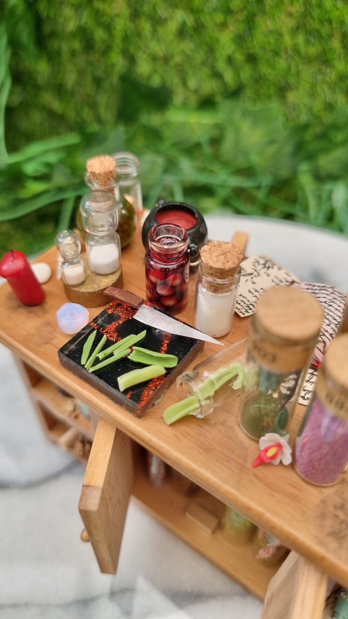 Top view of a 1:12 scale minature wooden sideboard, showing leaves on a black and red cutting board with knife, filled bottles, red candle, white flower, sandtimers, parchments and cauldron. The cupboard doors are open below