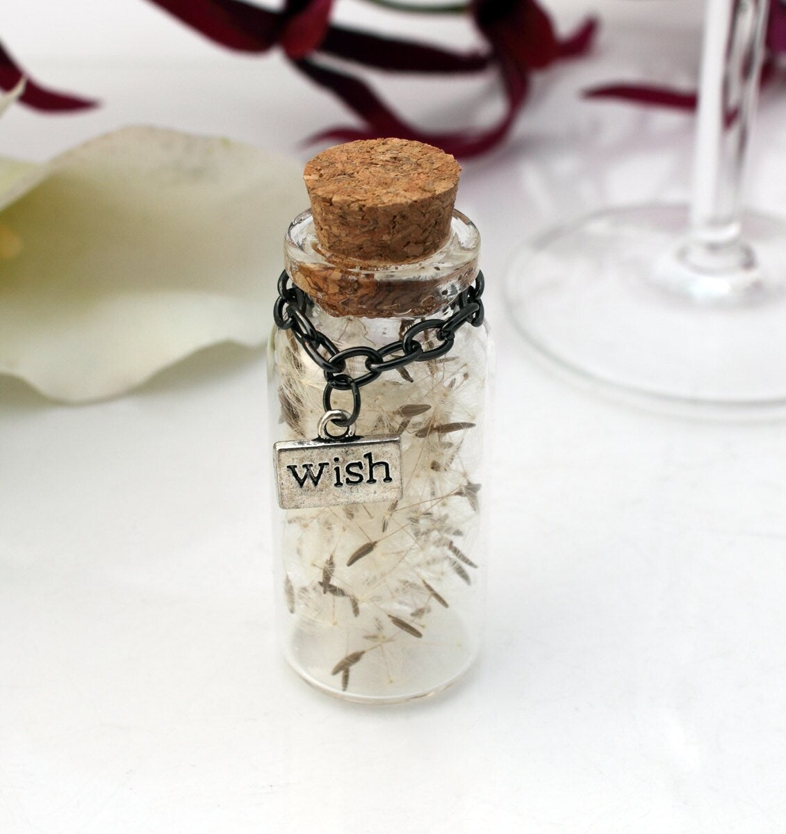 A single tall glass vial with cork lid containing dandelions seeds with silver coloured wish charm hanging from black chain around the jar necks. The jars are surrounded by paper confetti