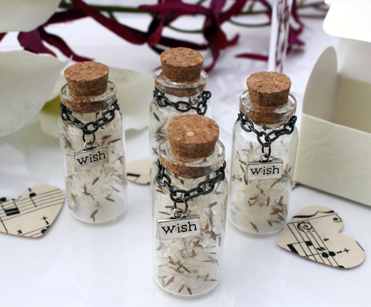 Four tall glass vials with cork lids containing dandelions seeds with silver coloured wish charms hanging from black chains around the jar necks. The jars are surrounded by paper confetti