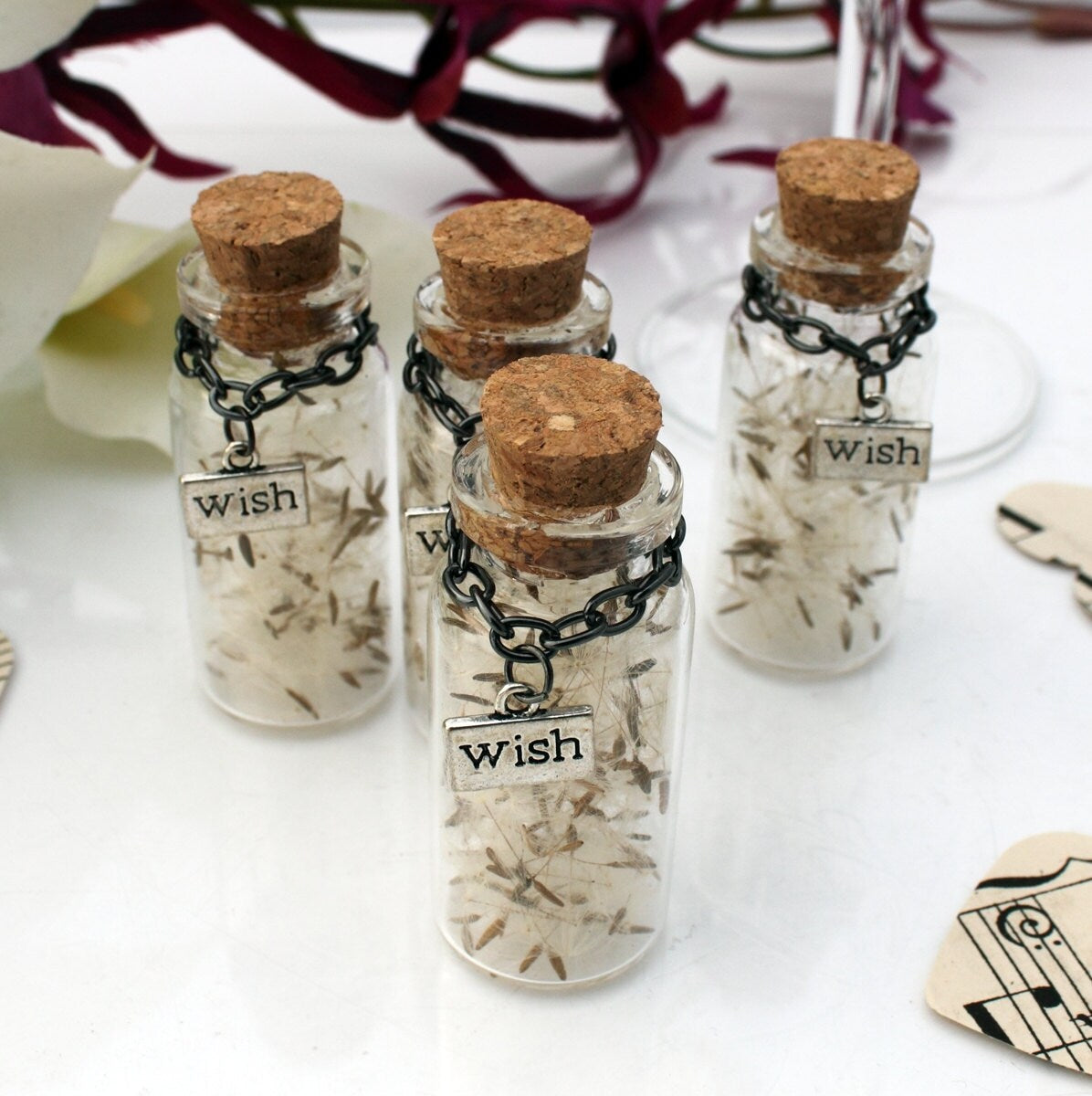 Four tall glass vials with cork lids containing dandelions seeds with silver coloured wish charms hanging from black chains around the jar necks. The jars are surrounded by paper confetti