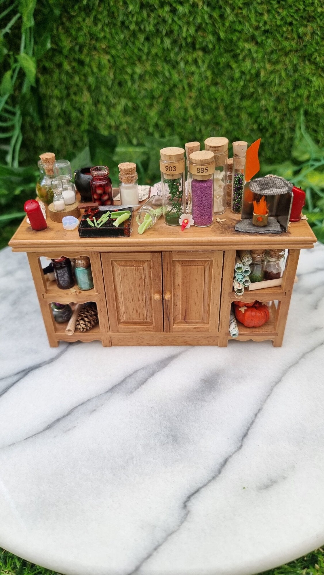 1:12 scale minature wooden sideboard with central cupboards and open shelves on the ends. The top is covered with parchment, wand, bottles of potion ingredients, open book, cauldron, notes, candles, sand timer, curios, inkwell and quill