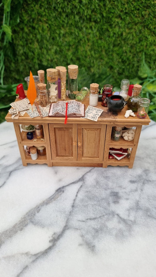 1:12 scale minature wooden sideboard with central cupboards and open shelves on the ends. The top is covered with parchment, wand, bottles of potion ingredients, open book, cauldron, notes, candles, sand timer, curios, inkwell and quill
