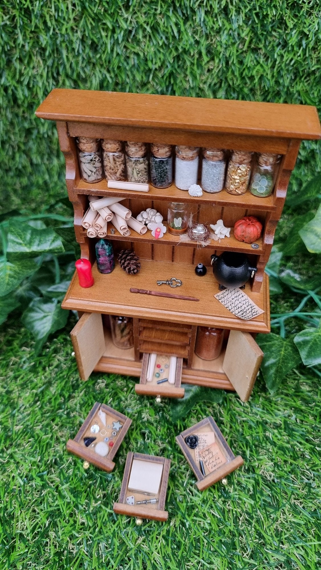 A 1:12 miniature brown dresser has 2 shelves with filled bottles, parchment rolls and curios. The counter top has a candle and cauldron. There are 2 open cupboard doors with bottles and 4 drawers laid in front notes and curios.
