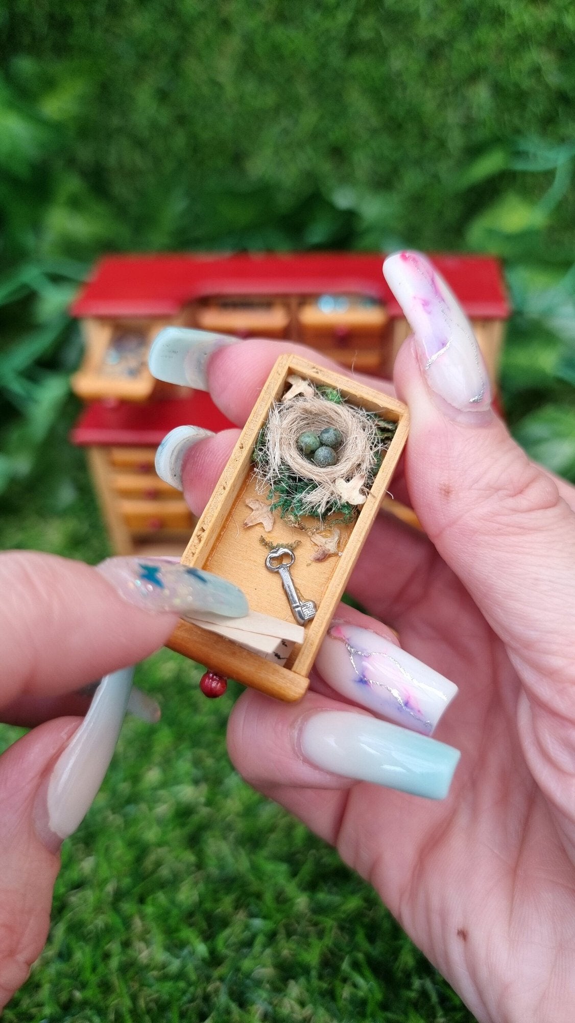 Hand holds a 1:12 drawer from a miniature desk. The drawer contains a tiny nest with 3 mottled green eggs, dead leaves and a key beneath a piece of parchment
