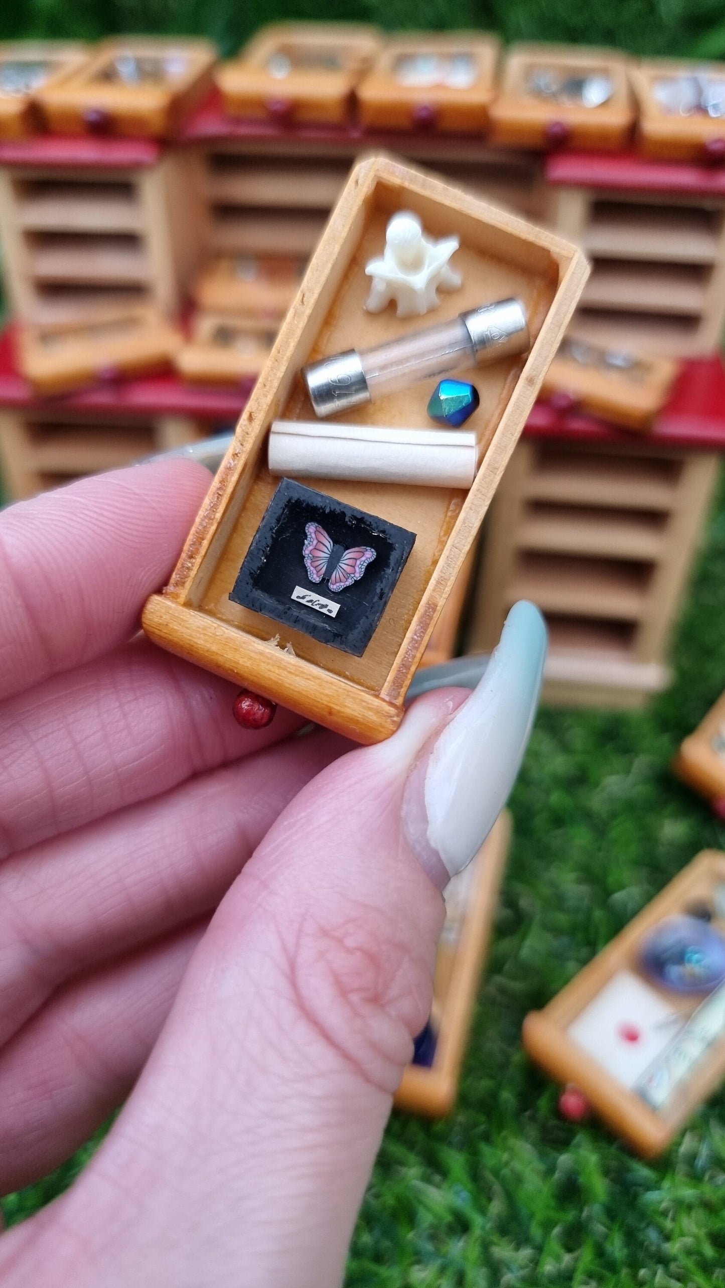 Hand holds a 1:12 drawer from a miniature desk. The drawer contains a bone, roll of parchment, glass tube and a tiny black butterfly shadow box with a pink and purple butterfly