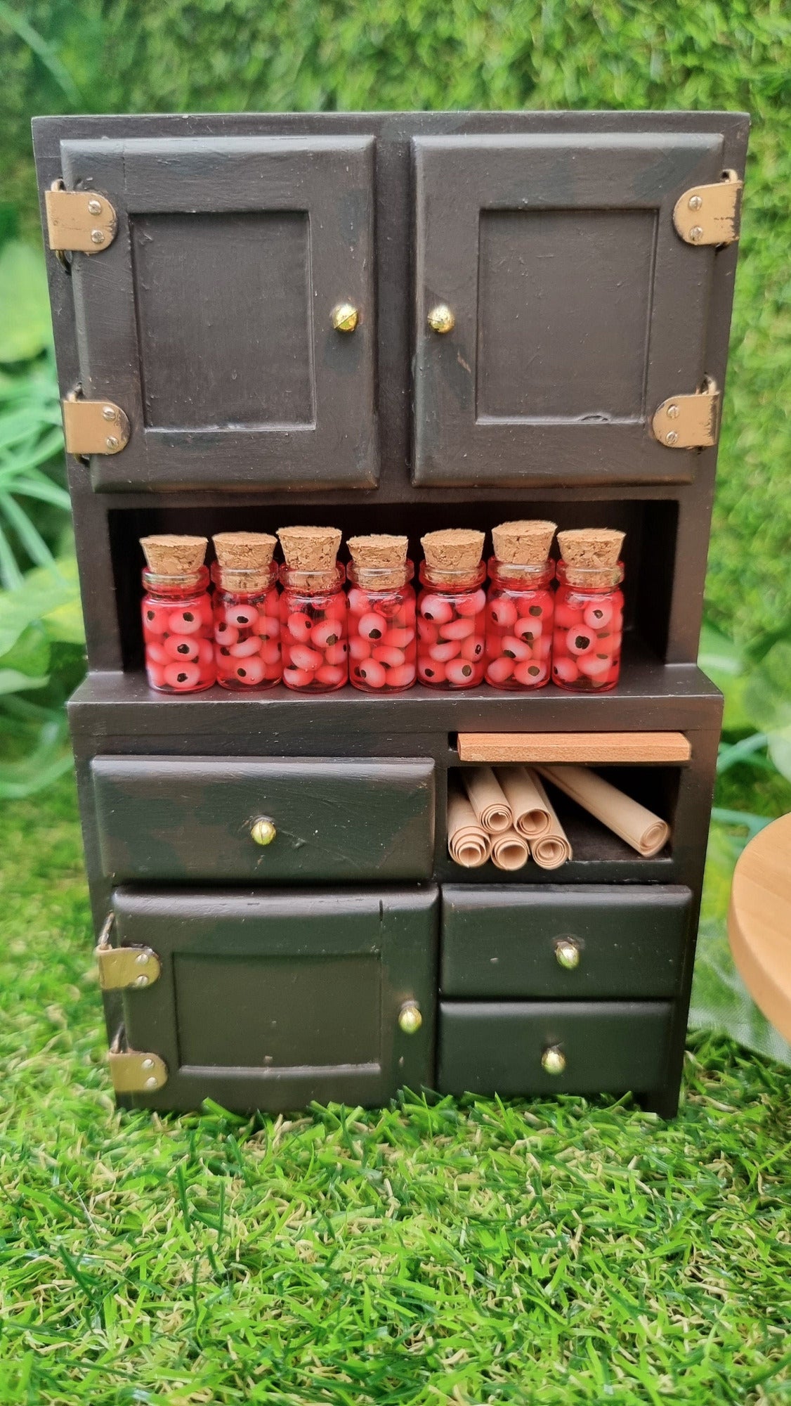 A 1:12 miniature black dresser holds 7 miniature glass jars containing eyeballs and red liquid. The miniature jars are topped with cork stoppers