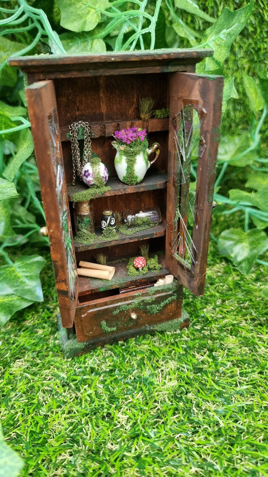 A miniature brown cabinet covered in moss sits open on a green background. The cabinet doors have shattered glass, and the mossy covered shelves hold vases, vessels, bottles, jug and a fly agaric mushroom. A bottom drawer sits slightly ajar