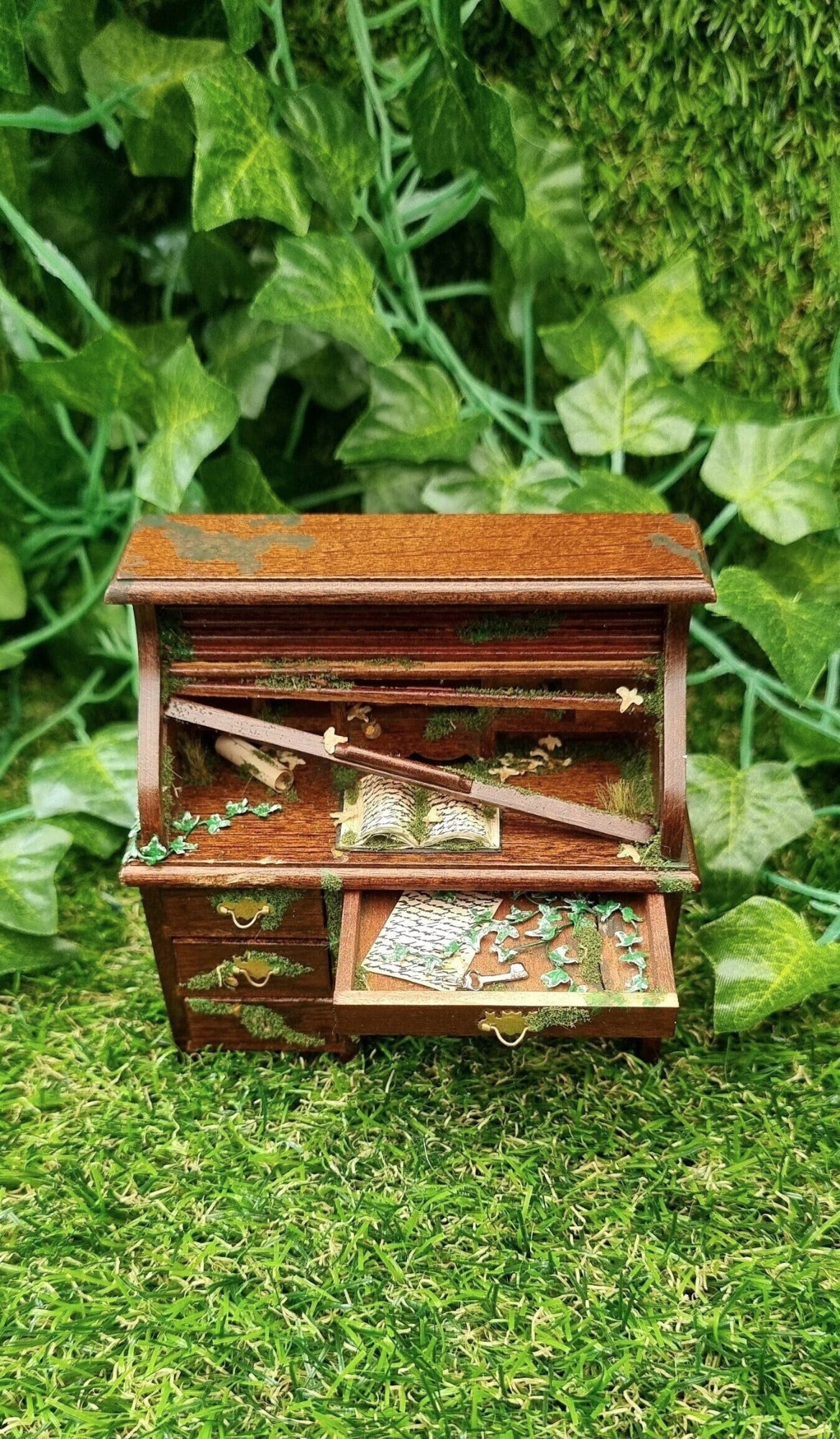 1:12 scale miniature abandoned rolltop desk with the top drawer open. The drawer contains green creeping ivy, moss, an old letter and a silver key. The desk top has a mossy old book, leaves and a broken wooden slat of the roll top