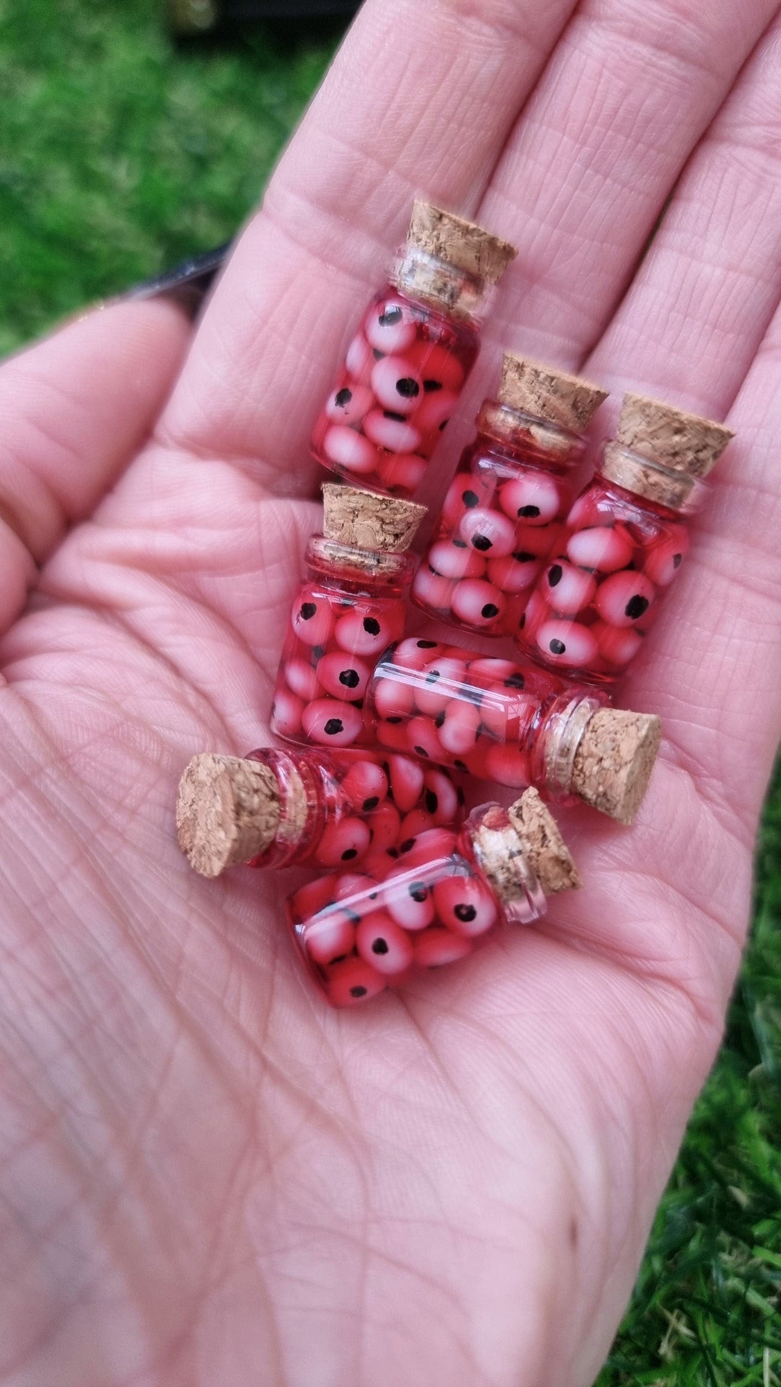 A hand holds 7 1:12 scale miniature glass jars containing eyeballs and red liquid. The miniature jars are topped with cork stoppers
