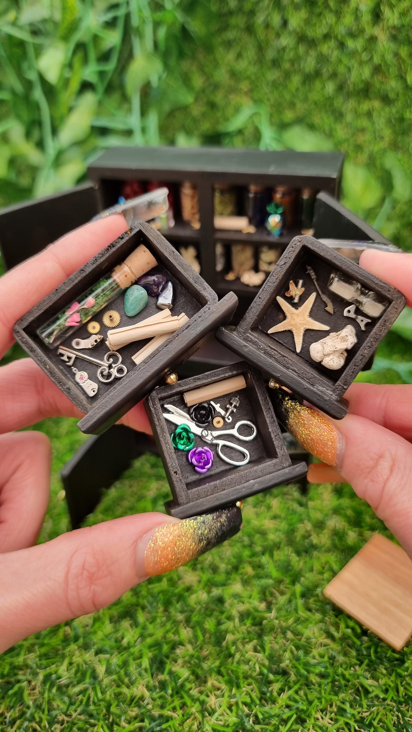 2 hands hold 3 1:12 scale miniature black drawers in front of a miniature dresser. The drawers contain flowers in a glass tube, key, parchments, starfish, coral, gemstones, sandtimer, scissors roses and trinkets