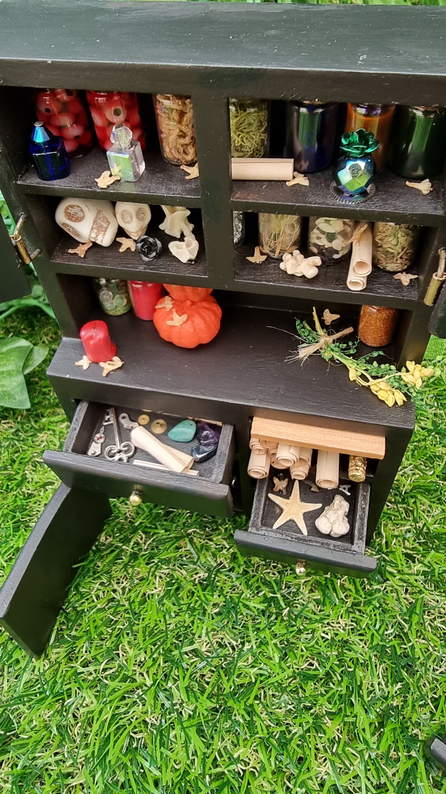 1:12 scale black dresser with open drawers and cupboards. The cupboards contain bottles of potion ingredients, curios, bone and skulls. The counter top has pumpkins, bottles, herbs and candle. The drawers contain parchment rolls, key and starfish
