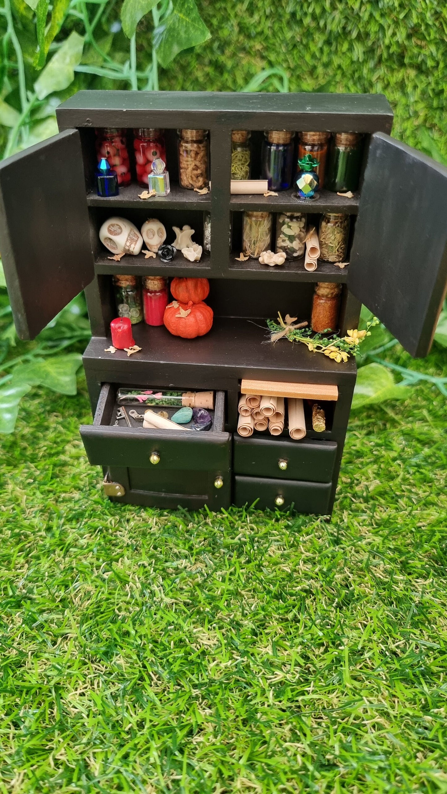 1:12 scale black dresser with open drawers and cupboards. The cupboards contain bottles of potion ingredients, curios, bone and skulls. The counter top has pumpkins bottles, herbs and candle. The drawers contain parchment rolls, key and starfish