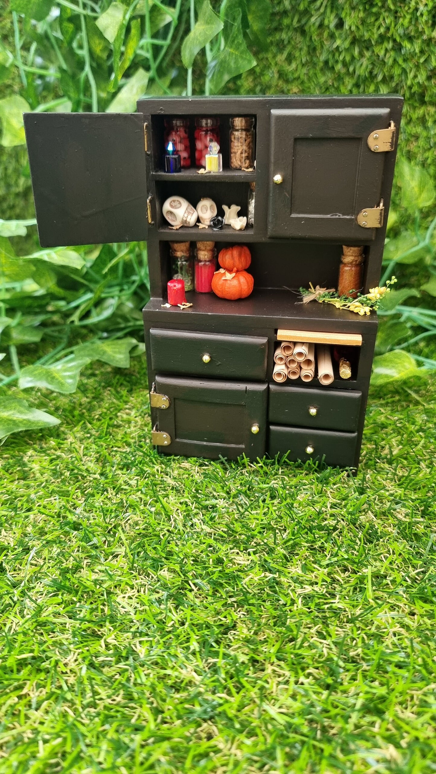 1:12 scale black dresser with an open cupboard containing bottles of potion ingredients, curios, bone and skulls. The counter top has pumpkins, bottles, herbs and candle. There is a nook filled with parchment rolls and 4 closed drawers