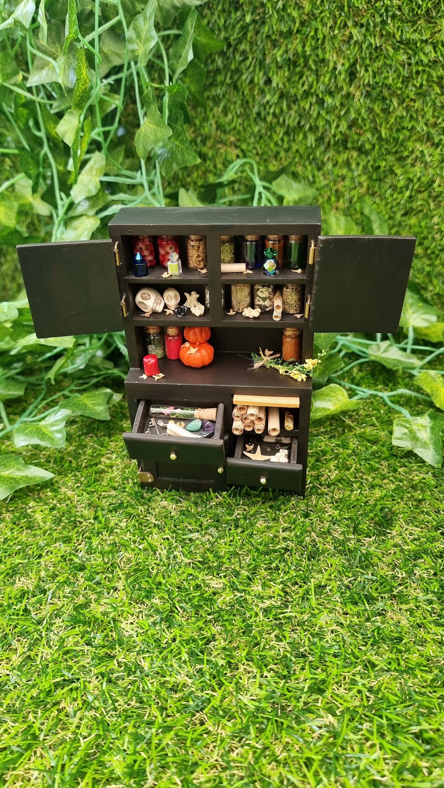 1:12 scale black dresser with open drawers and cupboards. They cupboards contains bottles of potion ingredients, curios, bone and skulls. The counter top has pumpkins bottles, herbs and candle. The drawers contain parchment rolls, key and starfish
