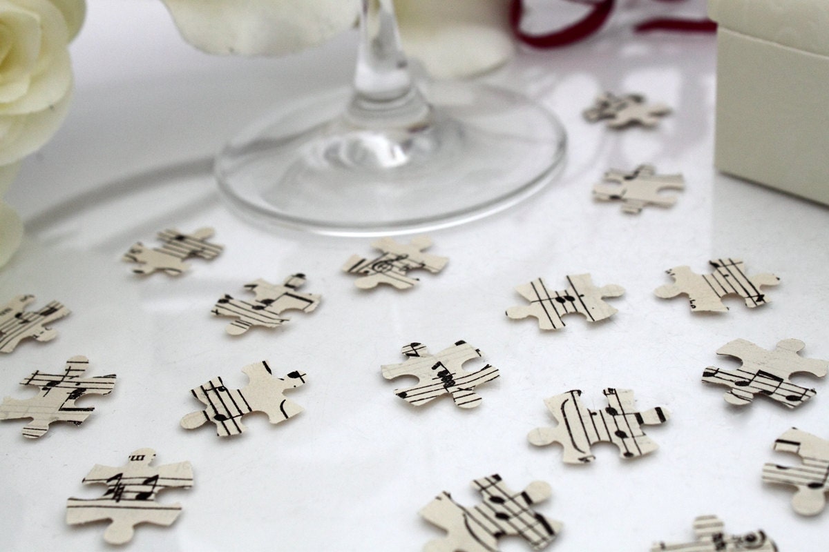 Cream paper confetti pieces in jigsaw shapes are scattered on a white background with a champagne flute and ivory rose orchid bouquet in the background. The paper confetti is cut from sheet music