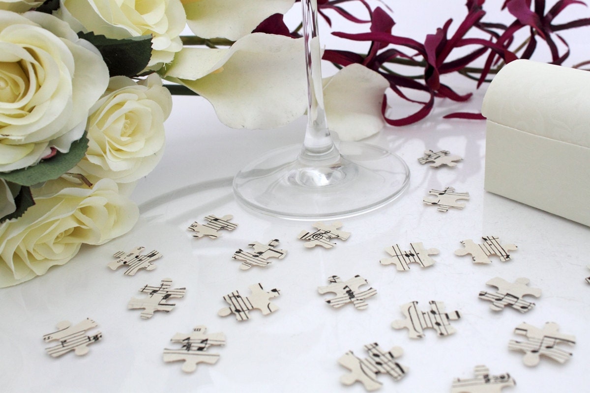 Cream paper confetti pieces in jigsaw shapes are scattered on a white background with a champagne flute and purple orchid bouquet in the background. The paper confetti is cut from sheet music