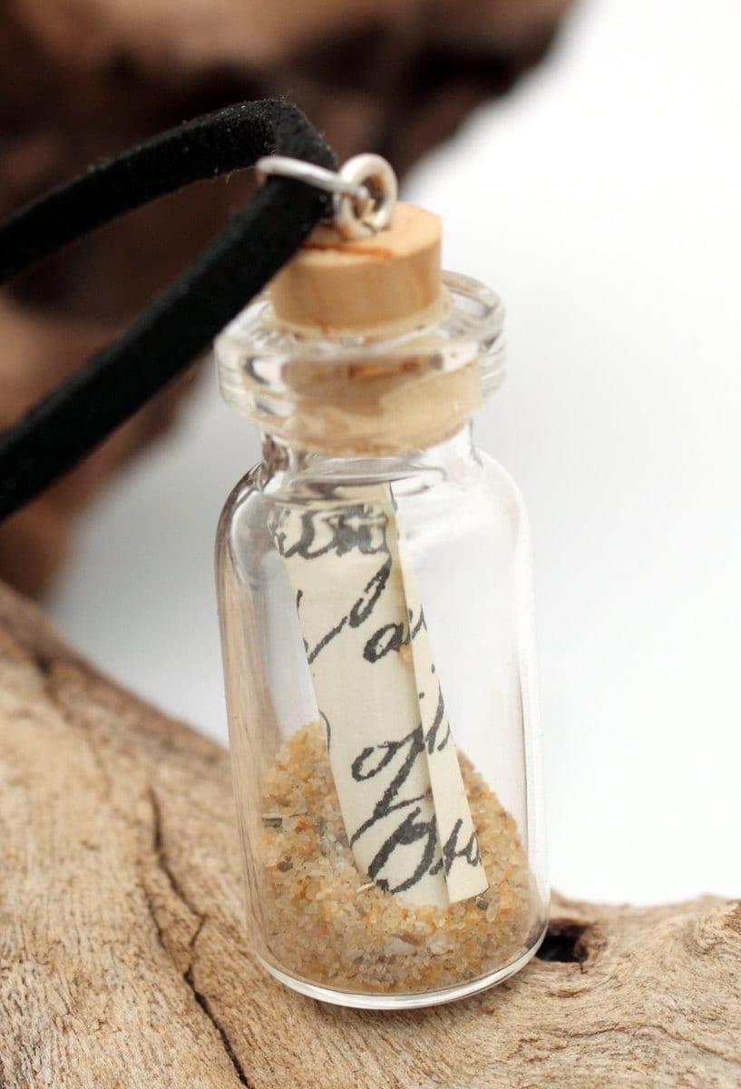 Small message in a bottle glass jar necklace containing a small scroll of paper and golden brown sand. The jar is displayed on a black coloured cord attached to the cork lid