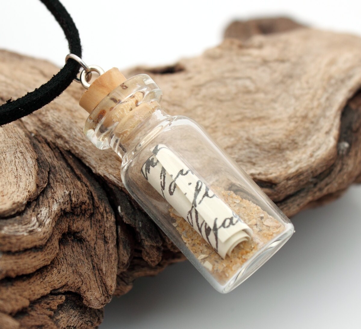 Small message in a bottle glass jar necklace containing a small scroll of paper and golden brown sand. The jar is displayed on a black coloured cord attached to the cork lid