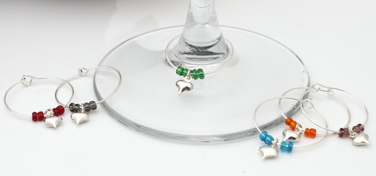 6 wine glass charms are arranged i beside a wine glass base on a white background. The wine glass charms are round wire hoops, each with different coloured seed beads and a silver coloured puffy heart. The green beaded charm is on the wine glass