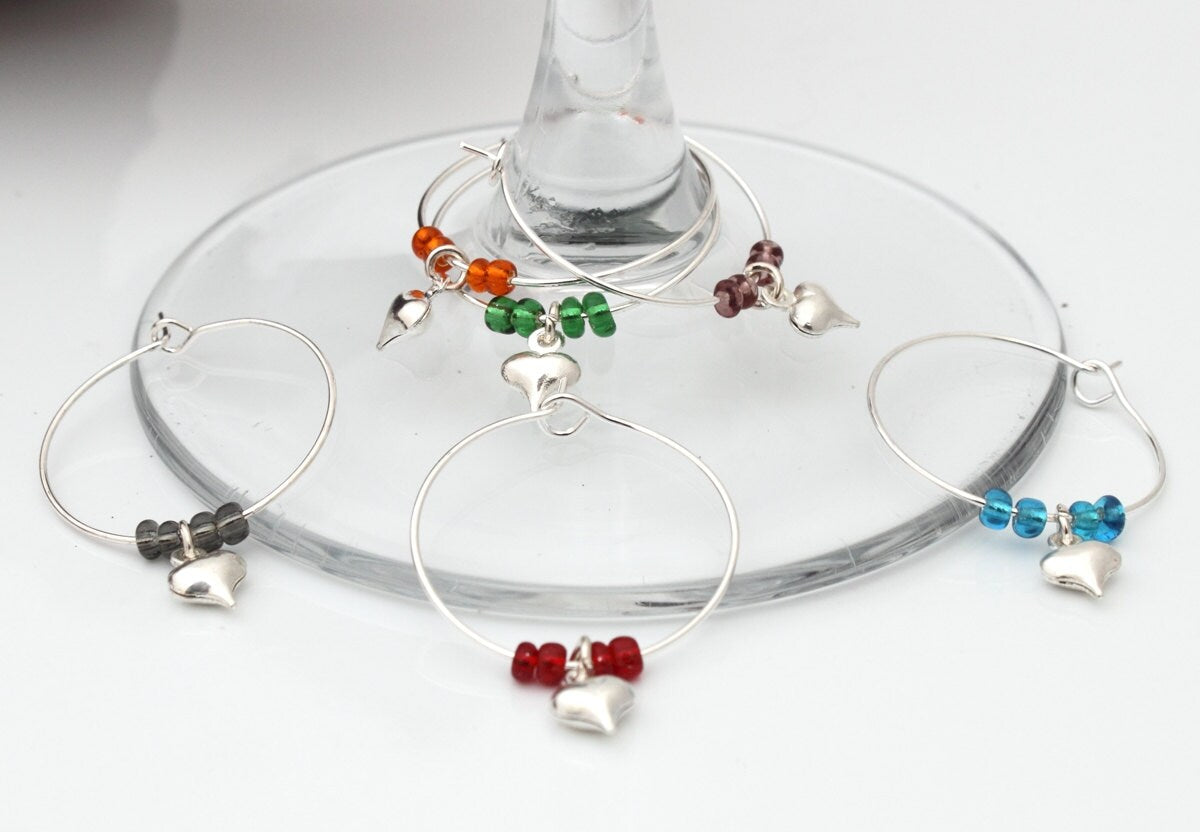 6 wine glass charms are arranged on top of a wine glass base on a white background. The wine glass charms are round wire hoops, each with different seed beads (orange, green, lavender, black red and turquoise) and a silver coloured puffy heart