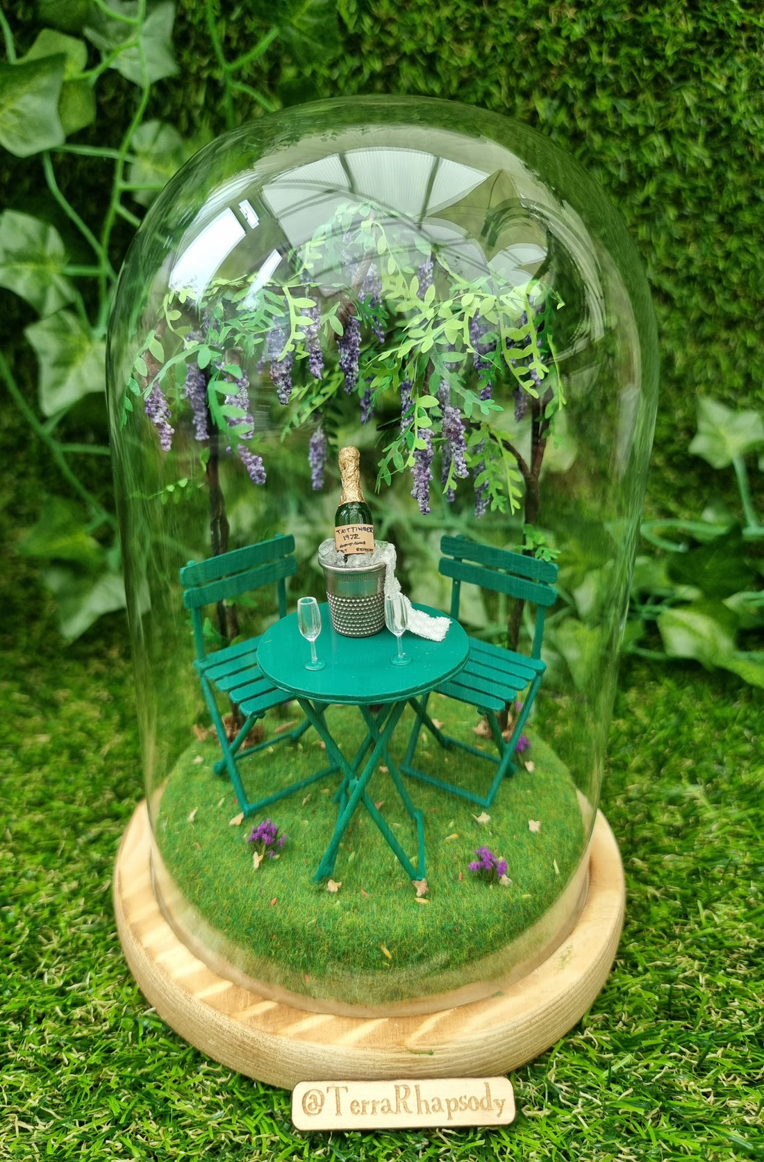 1:12 scale diorama under a glass bell jar, containing a miniature scene of a wisteria arch above a turquoise table and chairs with miniature champagne, champagne bucket and champagne flutes