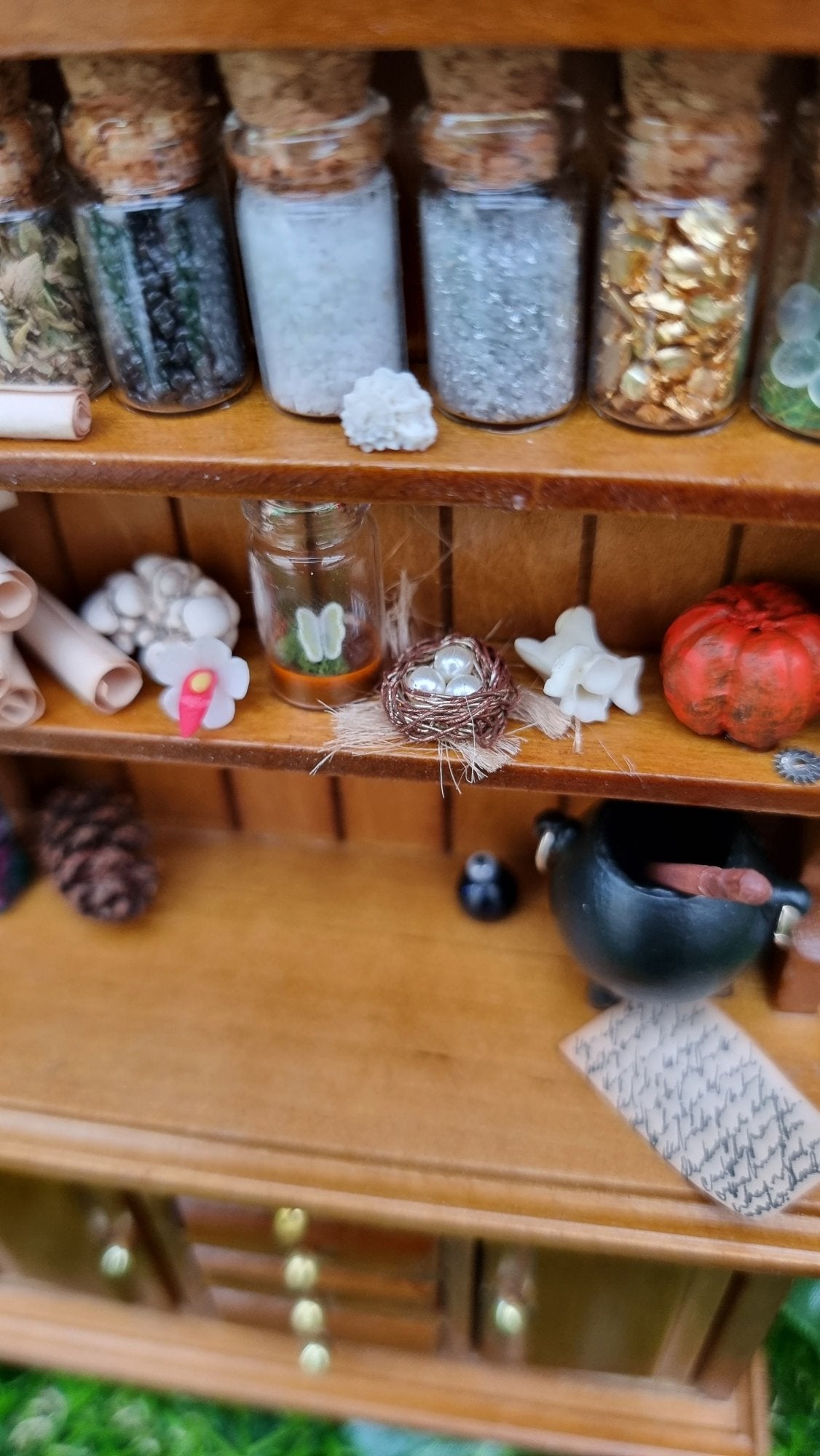 Detail of the 1:12 dresser is shown, with details of filled bottles, paper scrolls, butterfly bottle, birds nest, bone, pumpkion pine cone, written note and cauldron on the shelves and counter top.