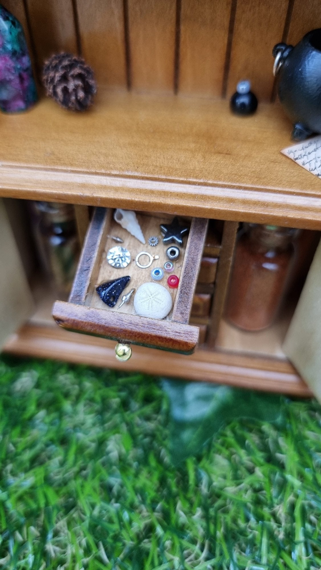 A 1:12 miniature brown dresser is shown with 1 open drawer with jewels, shells, sea urchin, curios and gemstones. Filled bottles, a pine cone and a cauldron can be seen in the background.