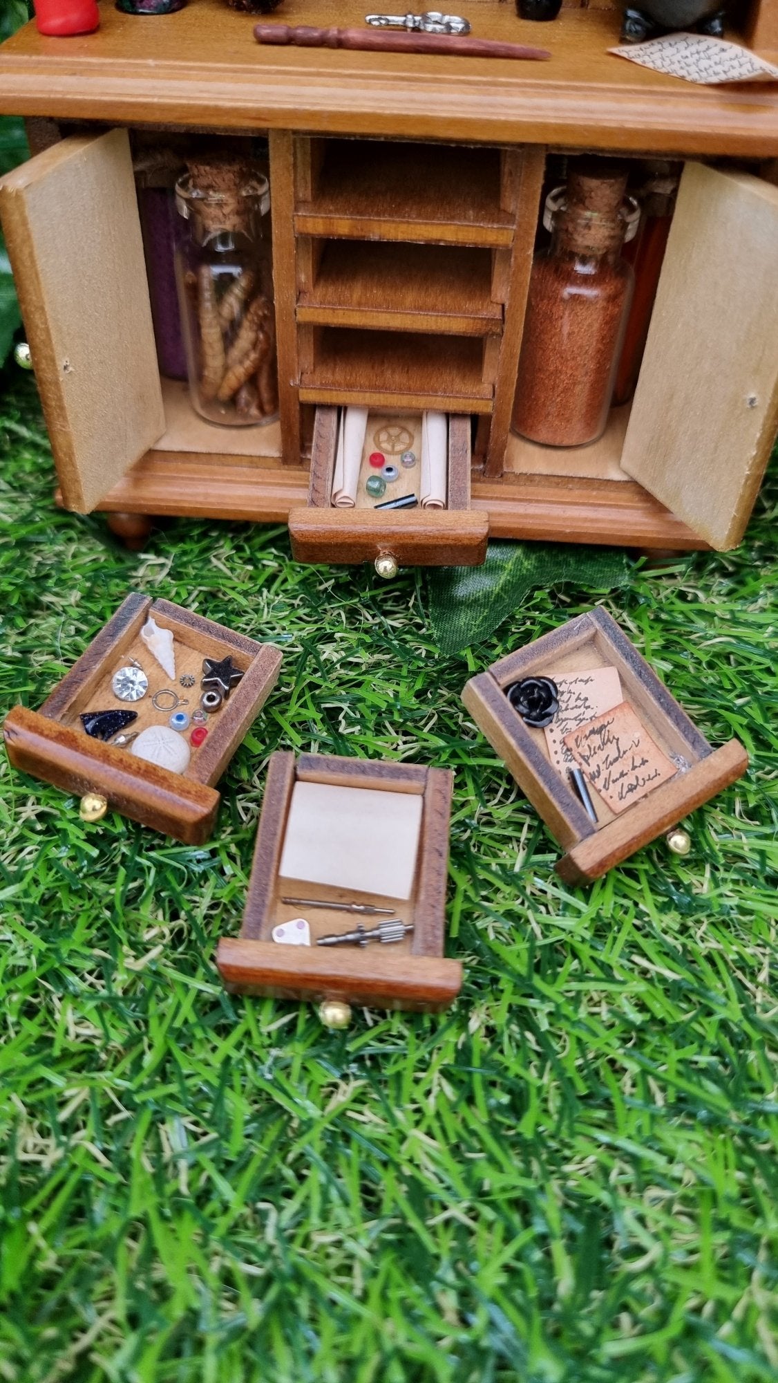 The bottom half of a 1:12 miniature brown dresser has 2 open cupboard doors with bottles and 4 drawers laid in front with a black rose, notes, scrolls and curios.