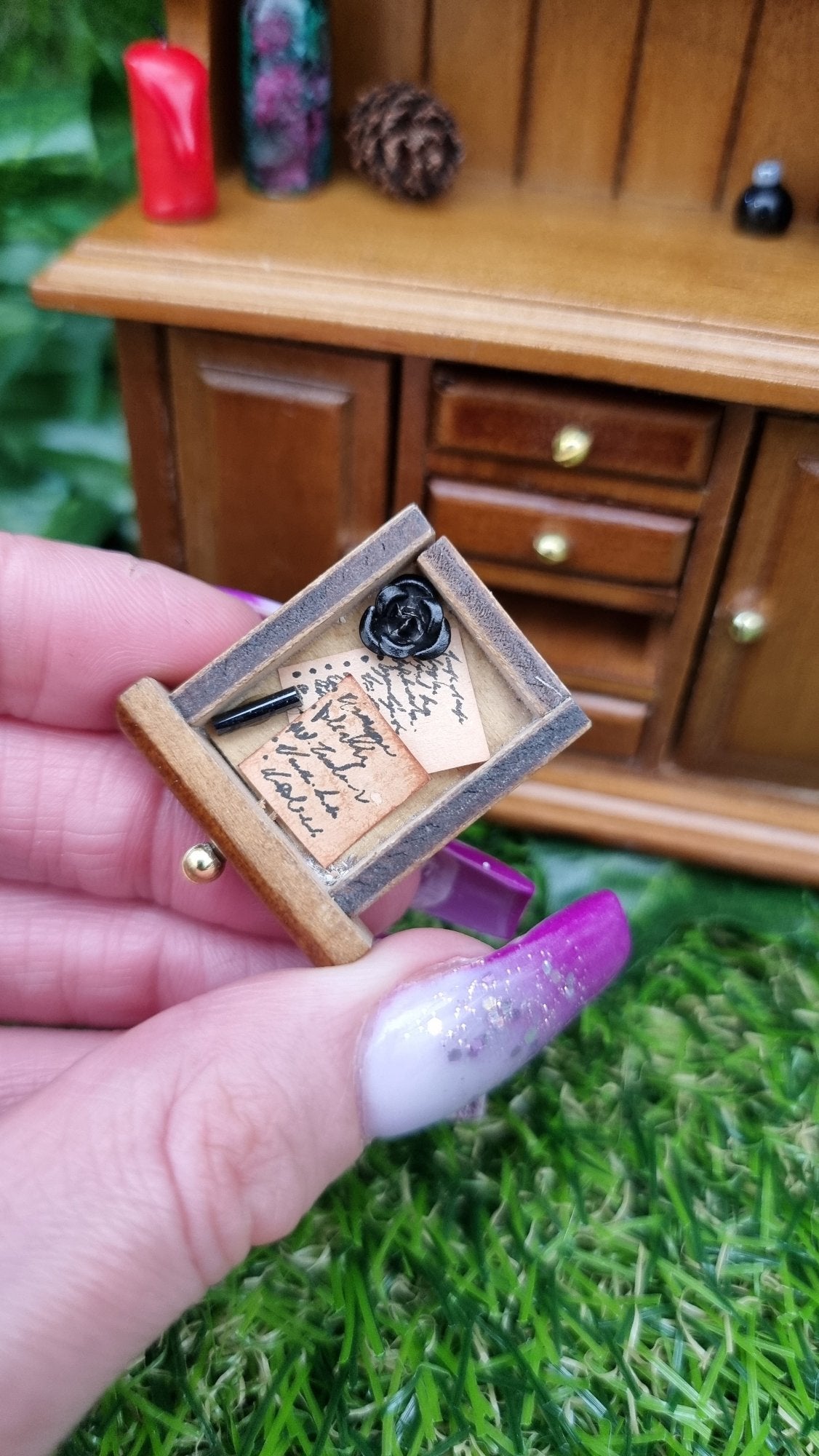 A miniature brown dresser is shown in the background while a hand holds a tiny drawer containing brown paper notes and a black metal rose. Ruby in zoisite, a pine cone and a red candle can be seen on the counter top