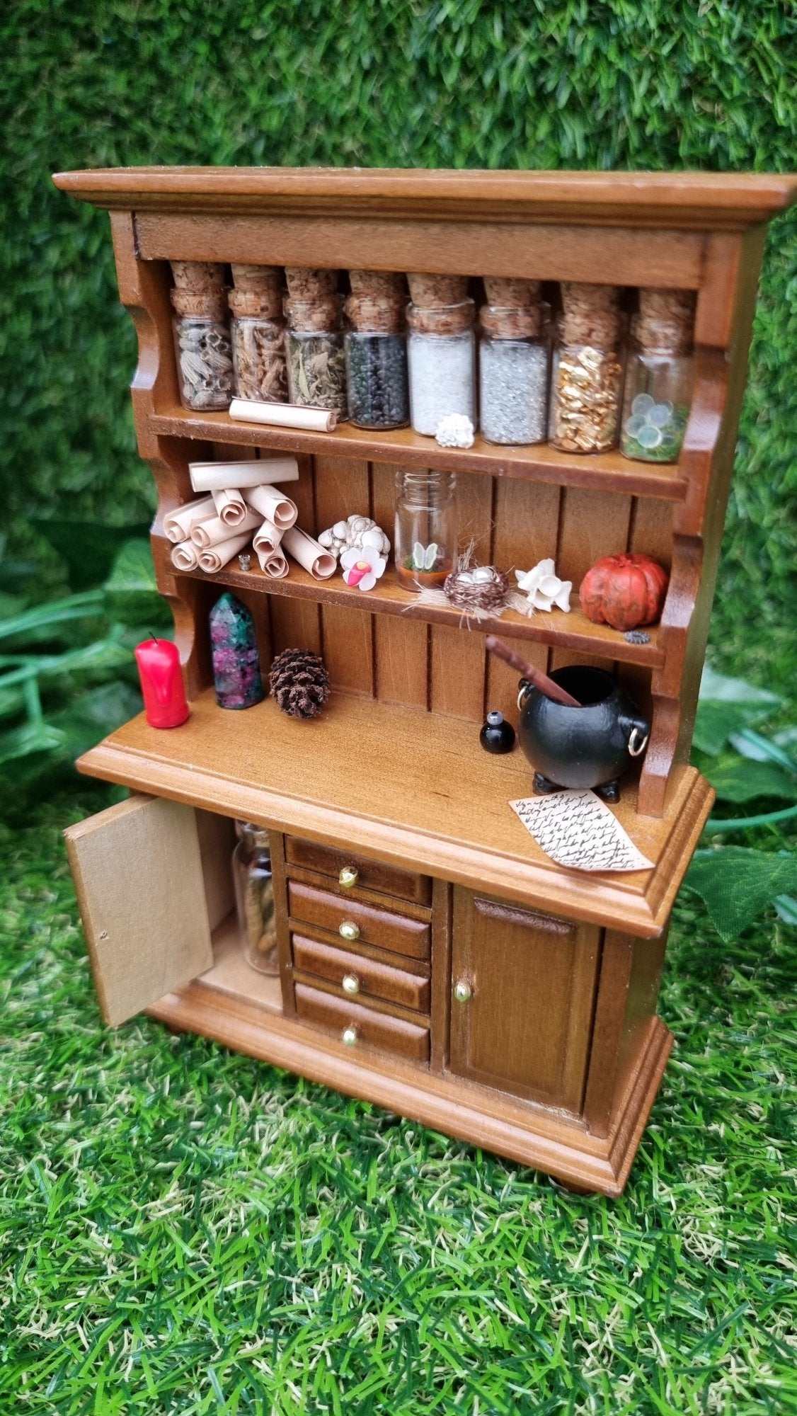 A 1:12 miniature brown dresser is shown against a green background. The dresser has 2 shelves with filled bottles, parchment rolls and curios. The counter top has a candle and cauldron. There are 2 cupboards with filled bottles and 4 drawers