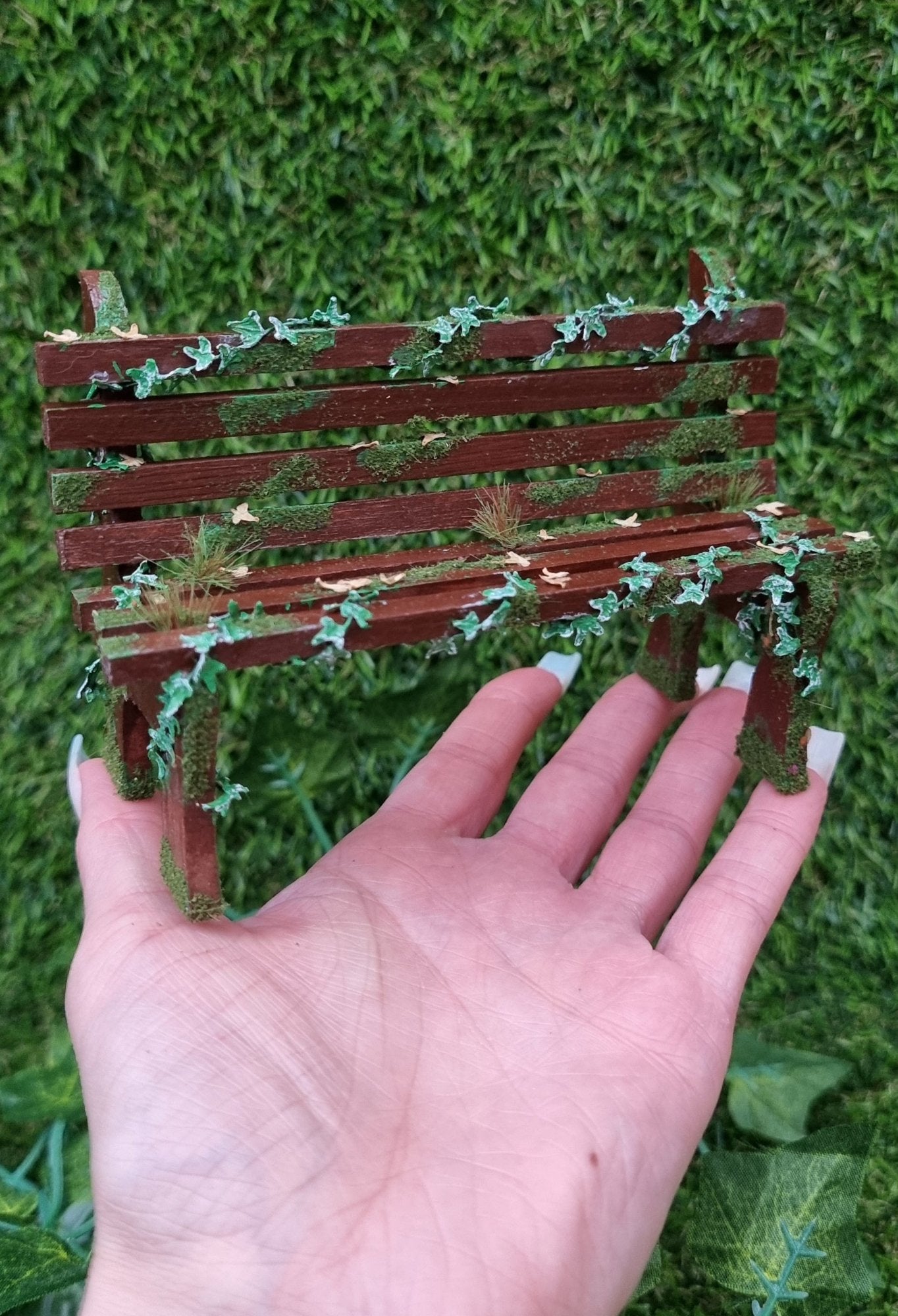 1:12 scale dollhouse wooden bench sits on a hand. The bench is abandoned, with green ivy twisting through the slats, dead leaves, grass and moss