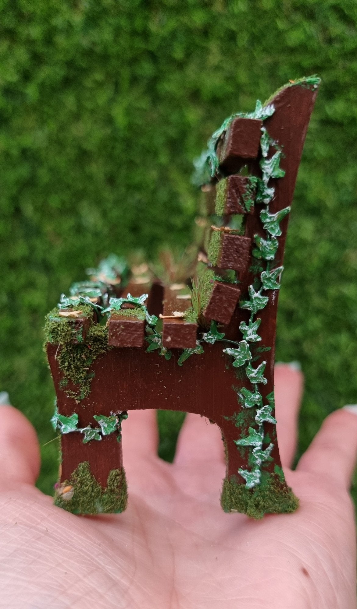 1:12 scale dollhouse wooden bench sits on a hand. The bench is abandoned, with green ivy twisting up the sides and through the slats, dead leaves, grass and moss