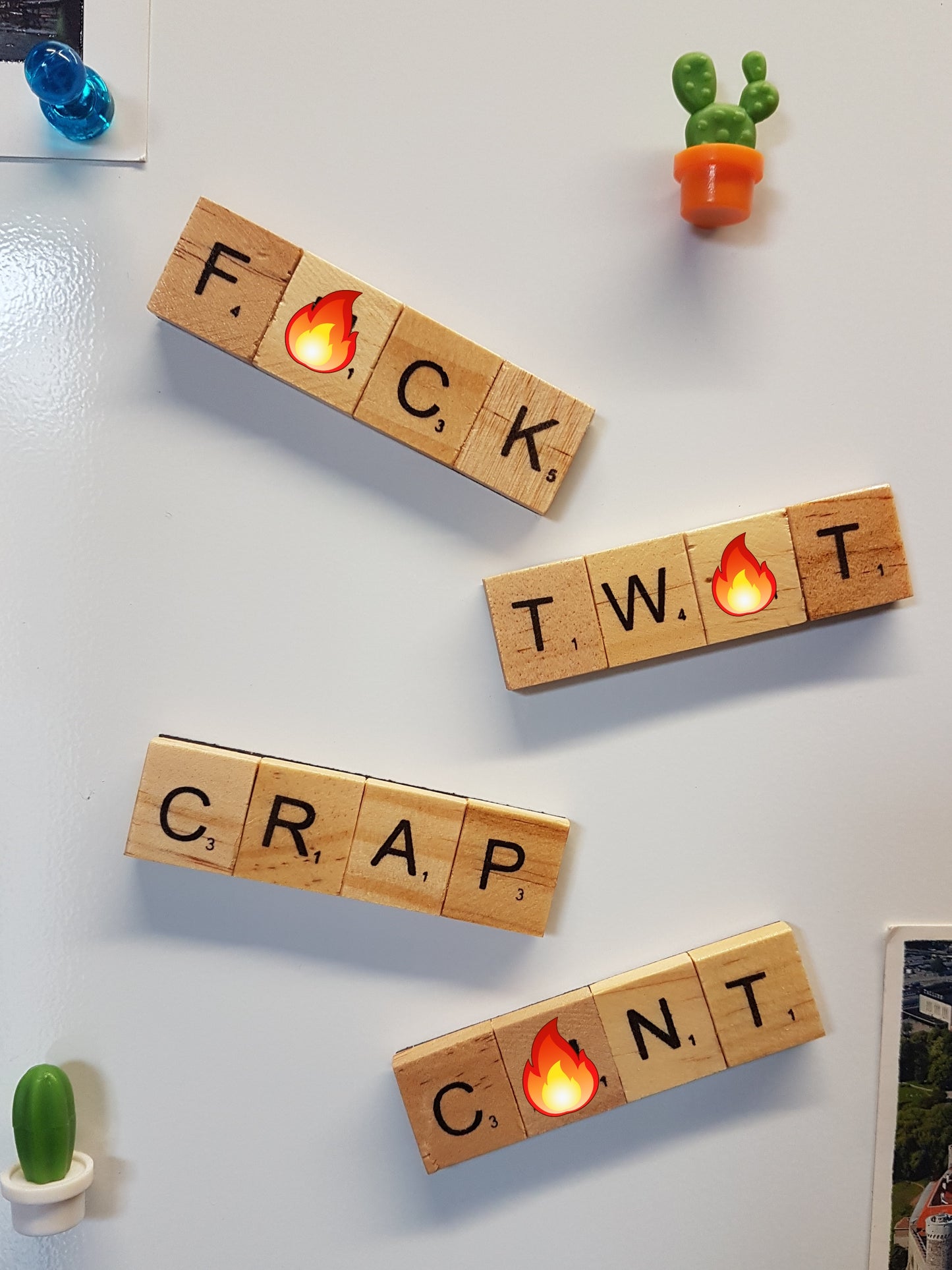 4 fridge magnets made with wooden scrabble tiles are stuck to the front of a fridge. The words spelled are swearwords, andeach swearword hasan emoji over one of the letters to read F#ck, Tw#t, Crap and C#nt