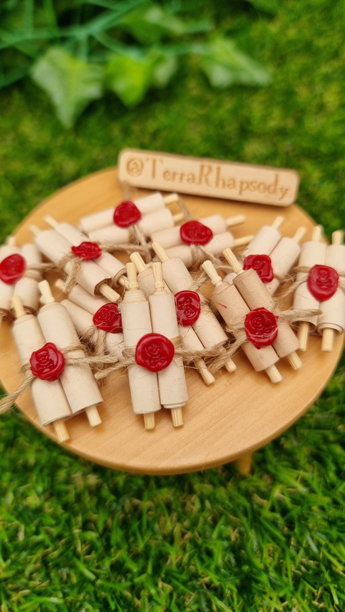 Miniature 1:12 scale double scrolls are arranged on a miniature wooden table. Each scroll has 2 rolls of paper around 2 wooden handles and bound with jute string a wax seal. The wax seal has a skull and crossbones indent. @TerraRhapsody is written on a sign in the background