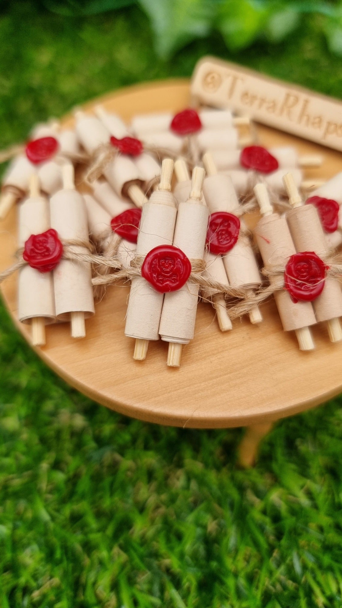 Miniature 1:12 scale double scrolls are arranged on a miniature wooden table. Each scroll has 2 rolls of paper around 2 wooden handles and bound with jute string a wax seal. The wax seal has a skull and crossbones indent. @TerraRhapsody is written on a sign in the background