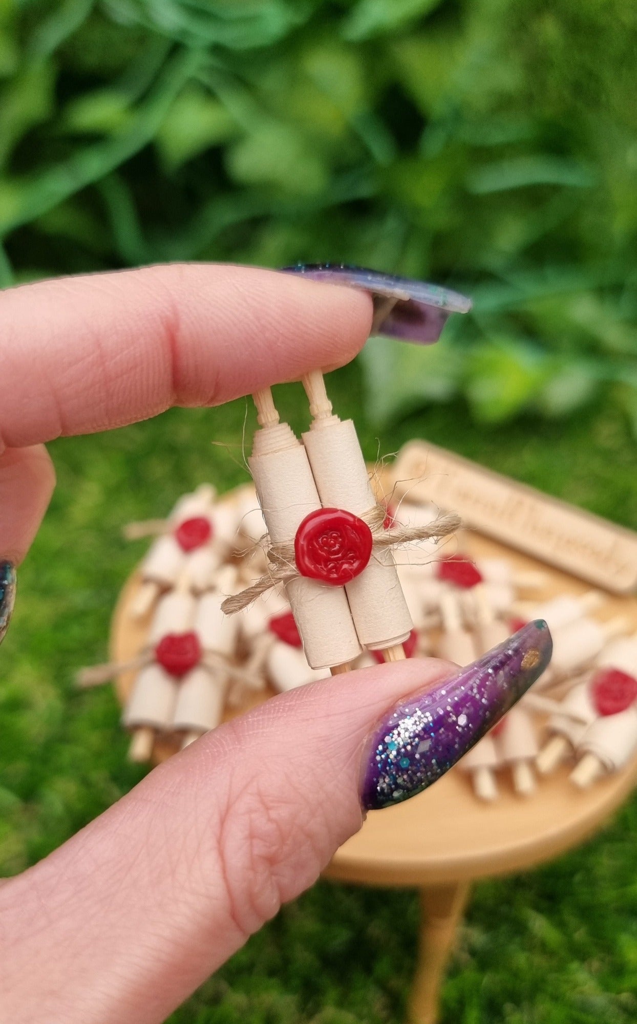 Miniature 1:12 scale double scroll is held between fingers in front of a miniature wooden table. The scroll has 2 rolls of paper around 2 wooden handles and bound with jute string a wax seal. The wax seal has a skull and crossbones indent. @TerraRhapsody is written on a sign in the background