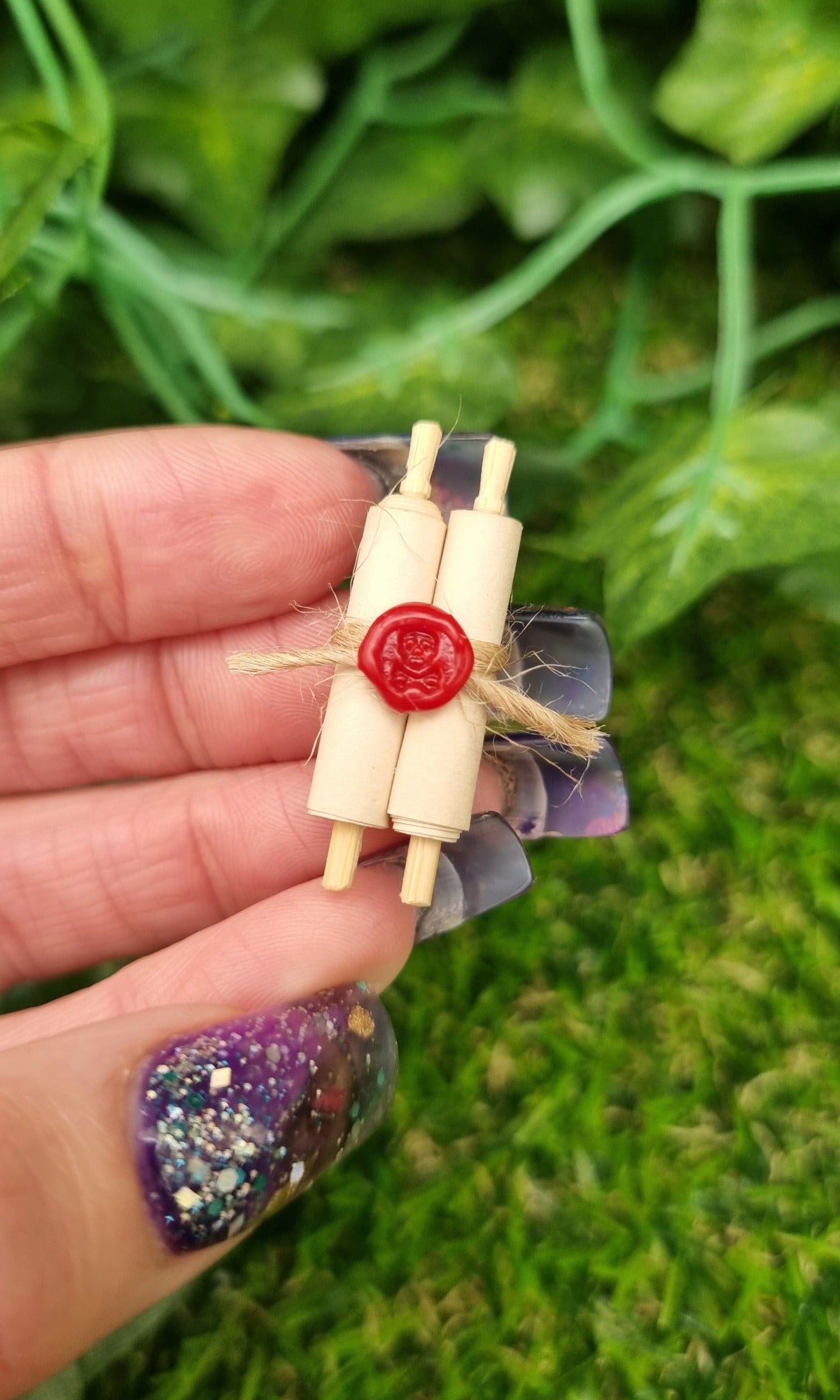 Miniature 1:12 scale double scroll is held between fingers. The scroll has 2 rolls of paper around 2 wooden handles and bound with jute string a wax seal. The wax seal has a skull and crossbones indent. 