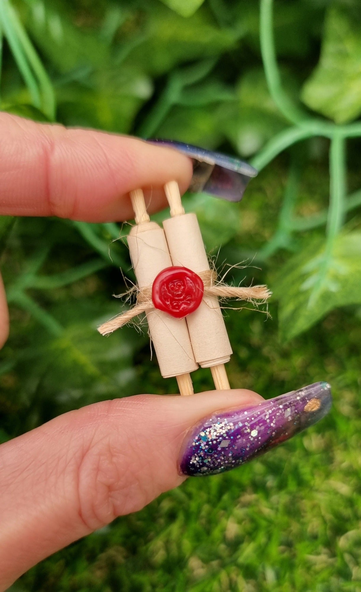 Miniature 1:12 scale double scroll is held between fingers.. The scroll has 2 rolls of paper around 2 wooden handles and bound with jute string a wax seal. The wax seal has a skull and crossbones indent.