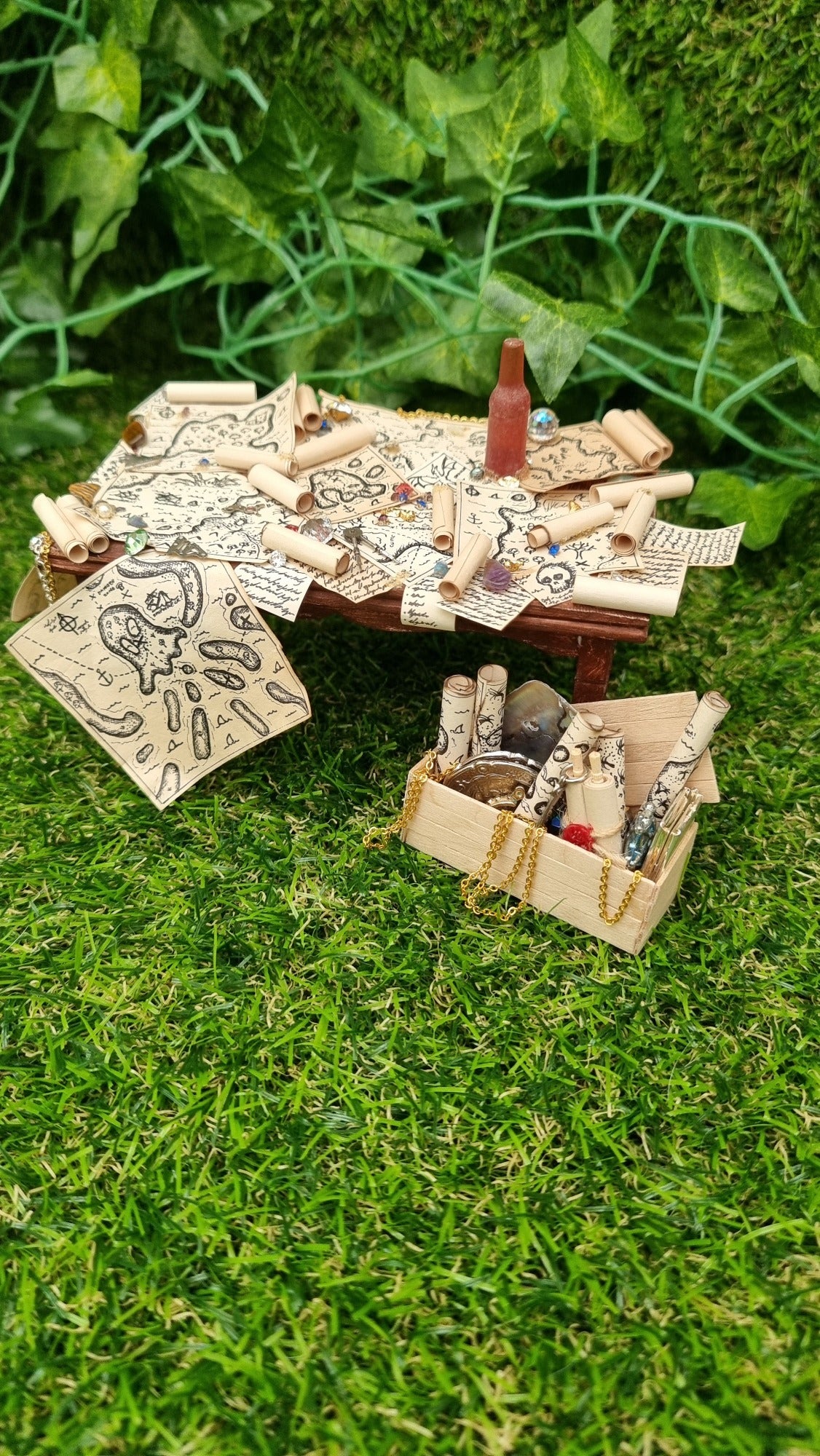 1:12 scale dollshouse brown table scattered with pirate treasure maps, bottle of rum and treasure. Wooden crate filled with treasure sits below the table